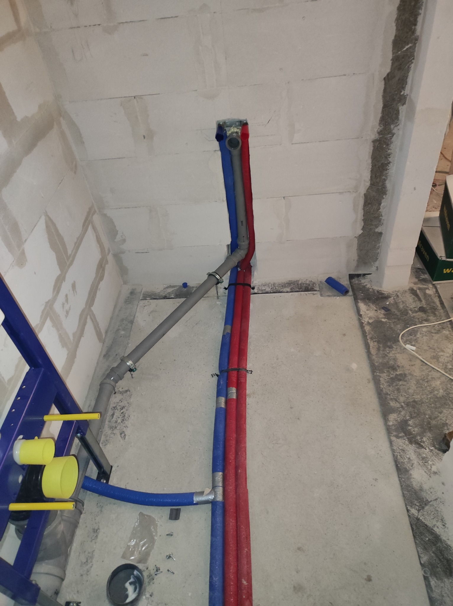 Surowe wnętrze łazienki w trakcie remontu, widoczne piony instalacji wodnej w kolorze czerwonym i niebieskim, szara rura kanalizacyjna oraz stelaż podtynkowy WC z żółtymi elementami.