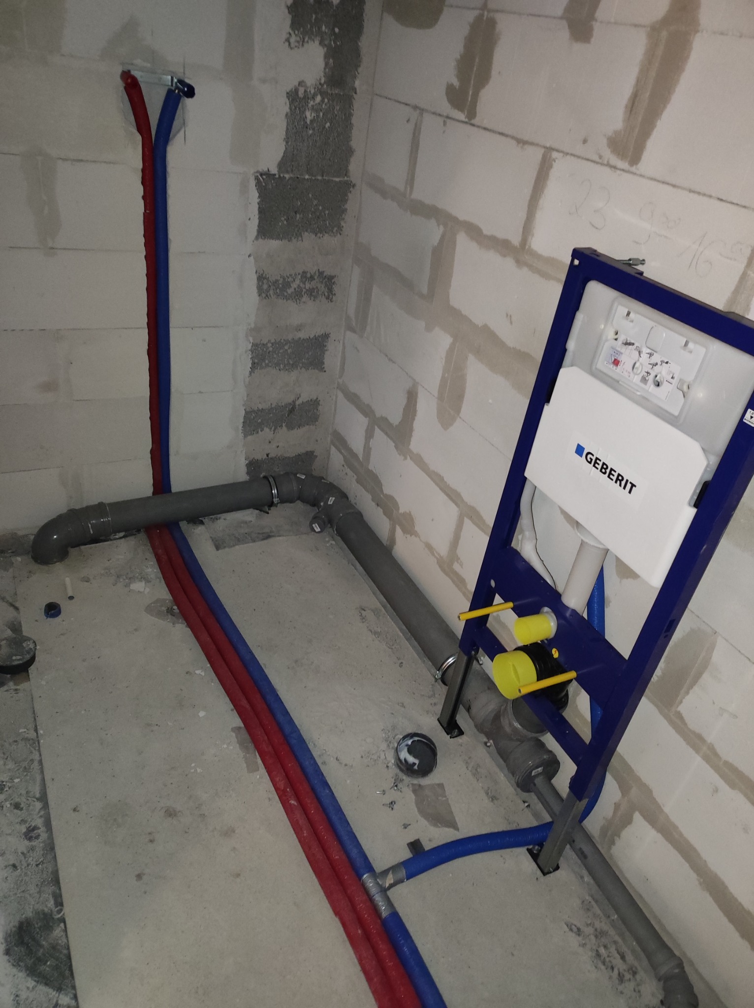 Surowy stan instalacji wodno-kanalizacyjnej w łazience z widocznym stelażem podtynkowym Geberit, rurami doprowadzającymi wodę oraz odpływami kanalizacyjnymi w pomieszczeniu z surowymi ścianami...