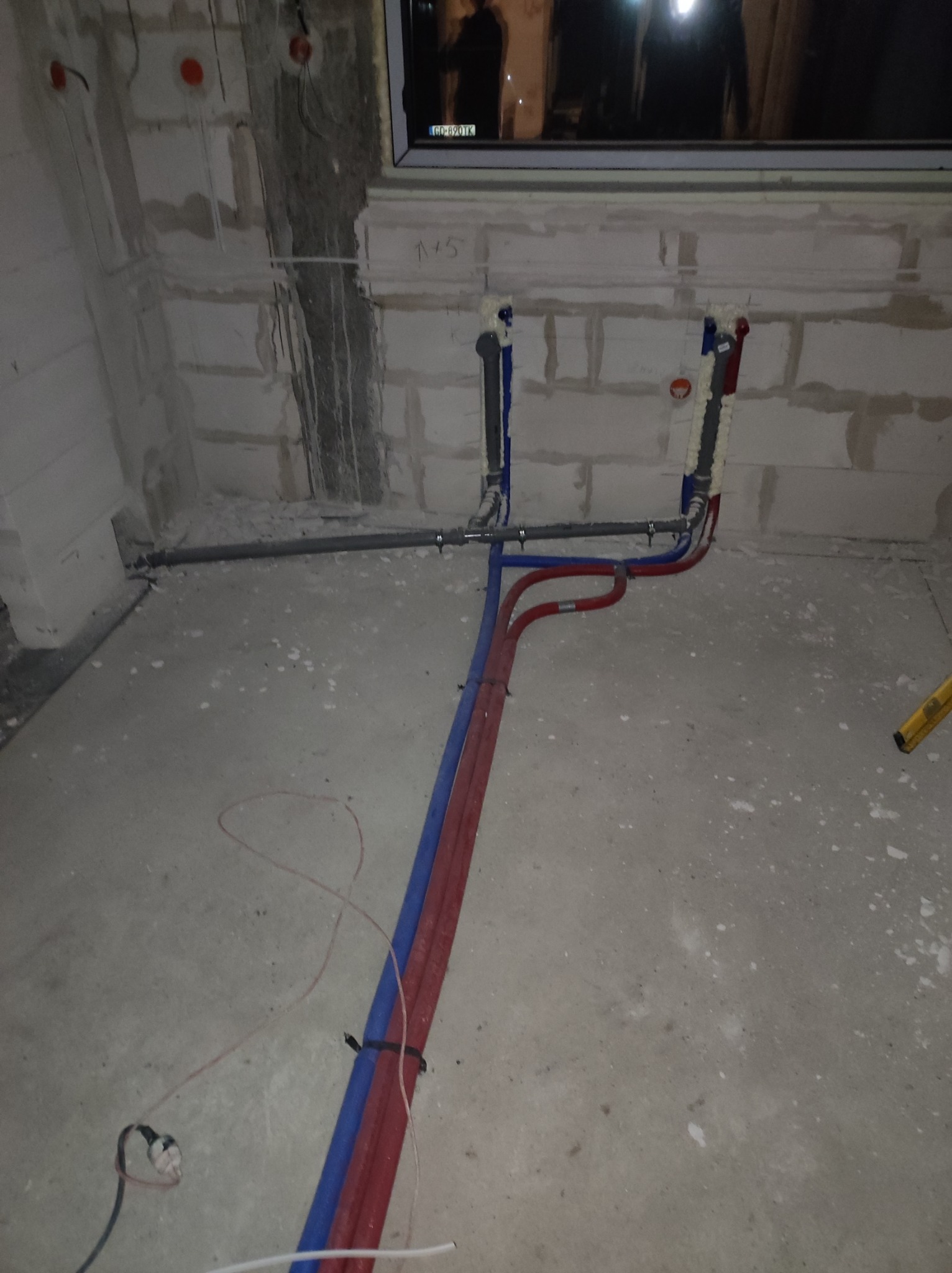 Surowe wnętrze w trakcie prac hydraulicznych: widoczne rury instalacji wodnej (czerwone i niebieskie) prowadzone po podłodze, przyłącza do ściany z pustaków, niezabezpieczone kable elektryczne...