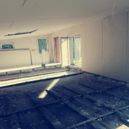 Wnętrze w trakcie remontu: goła podłoga z widoczną konstrukcją, ściany wykończone płytami gipsowo-kartonowymi, częściowo zamontowane okna i drzwi balkonowe.