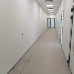 Długi korytarz biurowy z białymi, świeżo pomalowanymi ścianami i szarą podłogą z płytek. Widoczne niedociągnięcia w postaci kabli na podłodze oraz brakującej płyty w suficie podwieszanym.