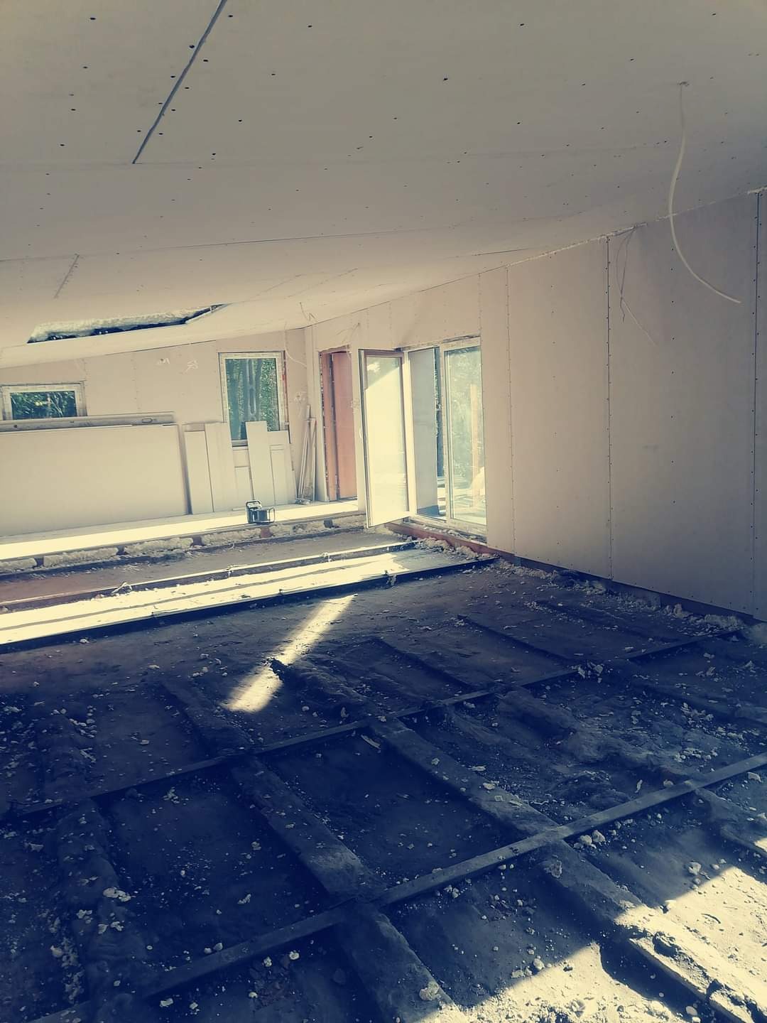 Wnętrze w trakcie remontu: goła podłoga z widoczną konstrukcją, ściany wykończone płytami gipsowo-kartonowymi, częściowo zamontowane okna i drzwi balkonowe.