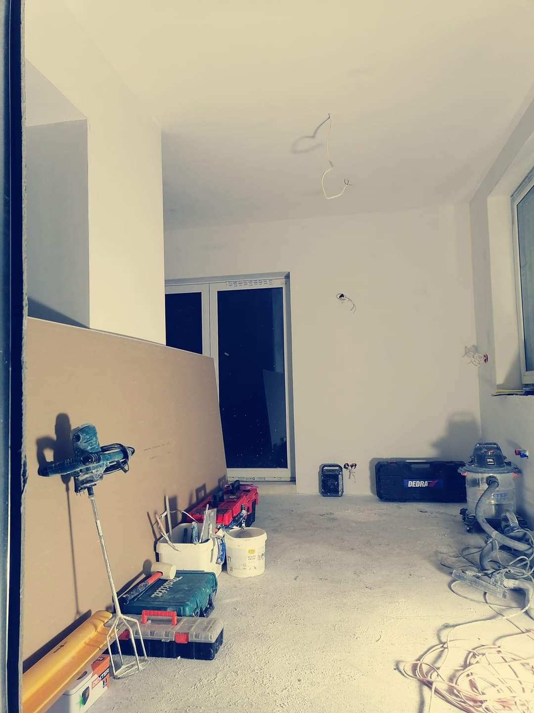 Wnętrze w trakcie remontu: płyta gipsowa oparta o ścianę, mikser do gładzi na stojaku, narzędzia w kuwetach, odkurzacz przemysłowy, surowe ściany przygotowane do wykończenia, wiszące przewody...