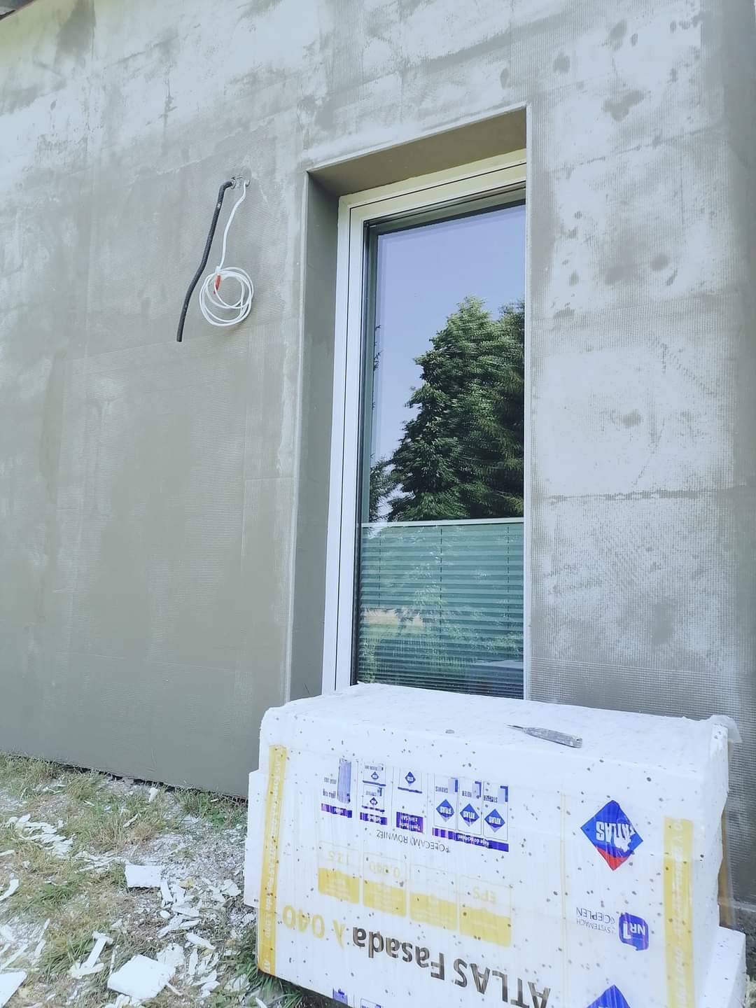 Świeżo otynkowana elewacja budynku jednorodzinnego, widoczna warstwa siatki, okno z roletami, paczka styropianu fasadowego ATLAS i resztki styropianu na trawie.