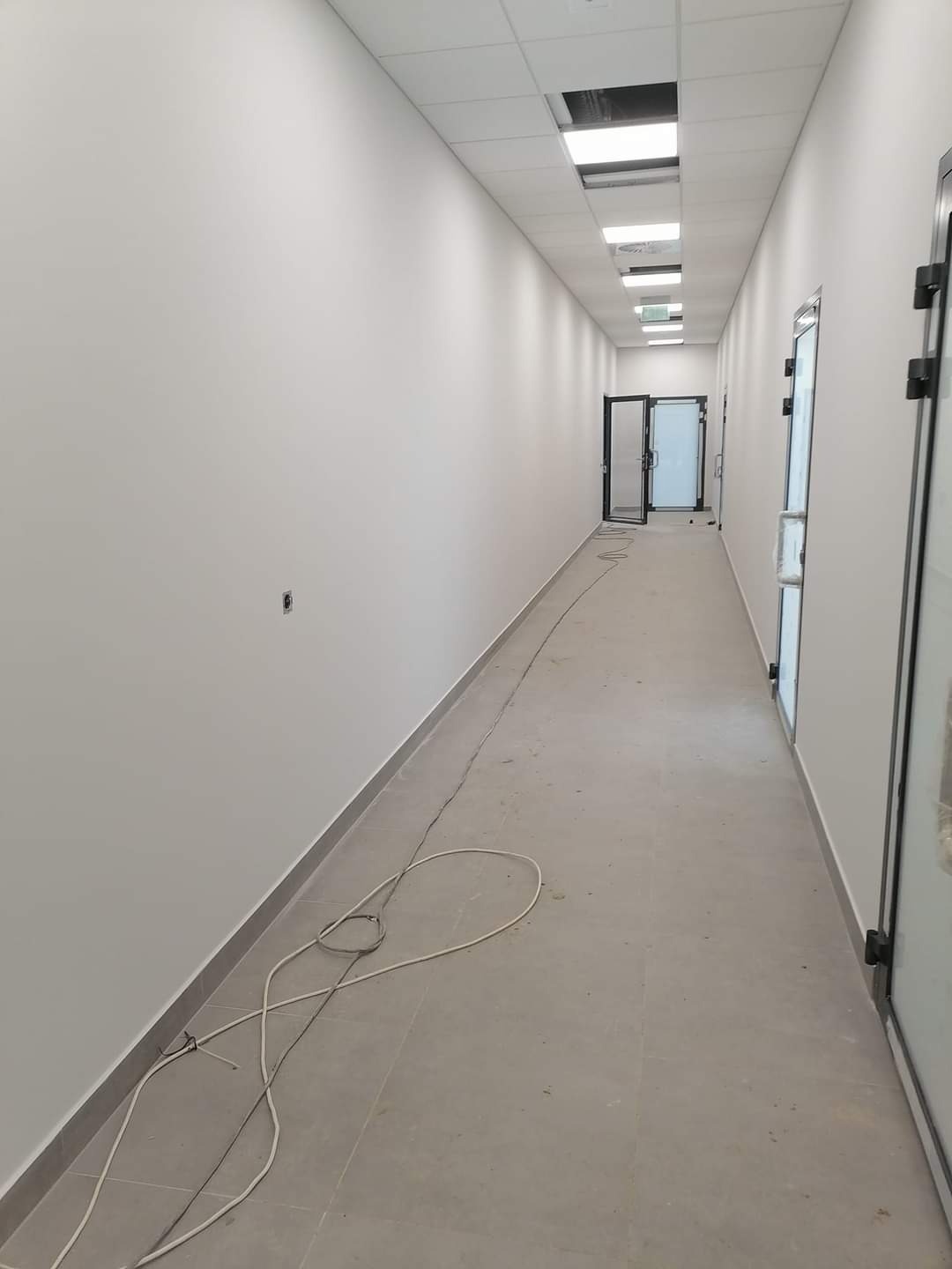 Długi korytarz biurowy z białymi, świeżo pomalowanymi ścianami i szarą podłogą z płytek. Widoczne niedociągnięcia w postaci kabli na podłodze oraz brakującej płyty w suficie podwieszanym.