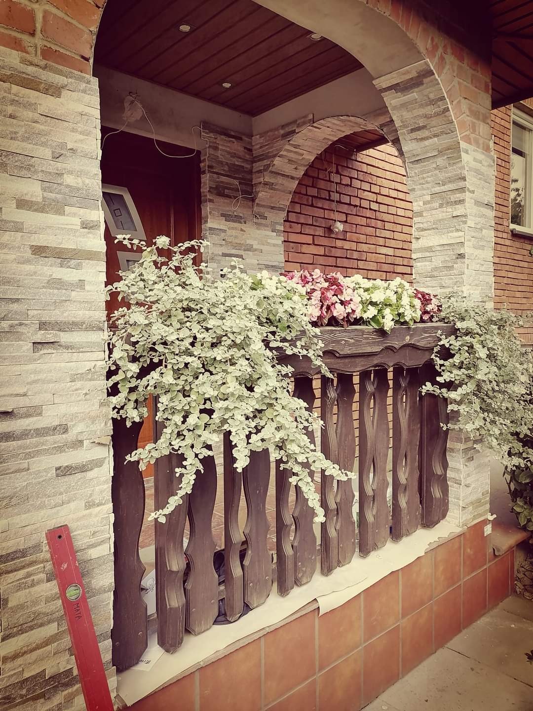 Wejście do domu z łukowym portykiem wykończonym kamieniem elewacyjnym, drewnianą balustradą zdobioną kwiatami i czerwoną poziomicą opartą o ścianę.