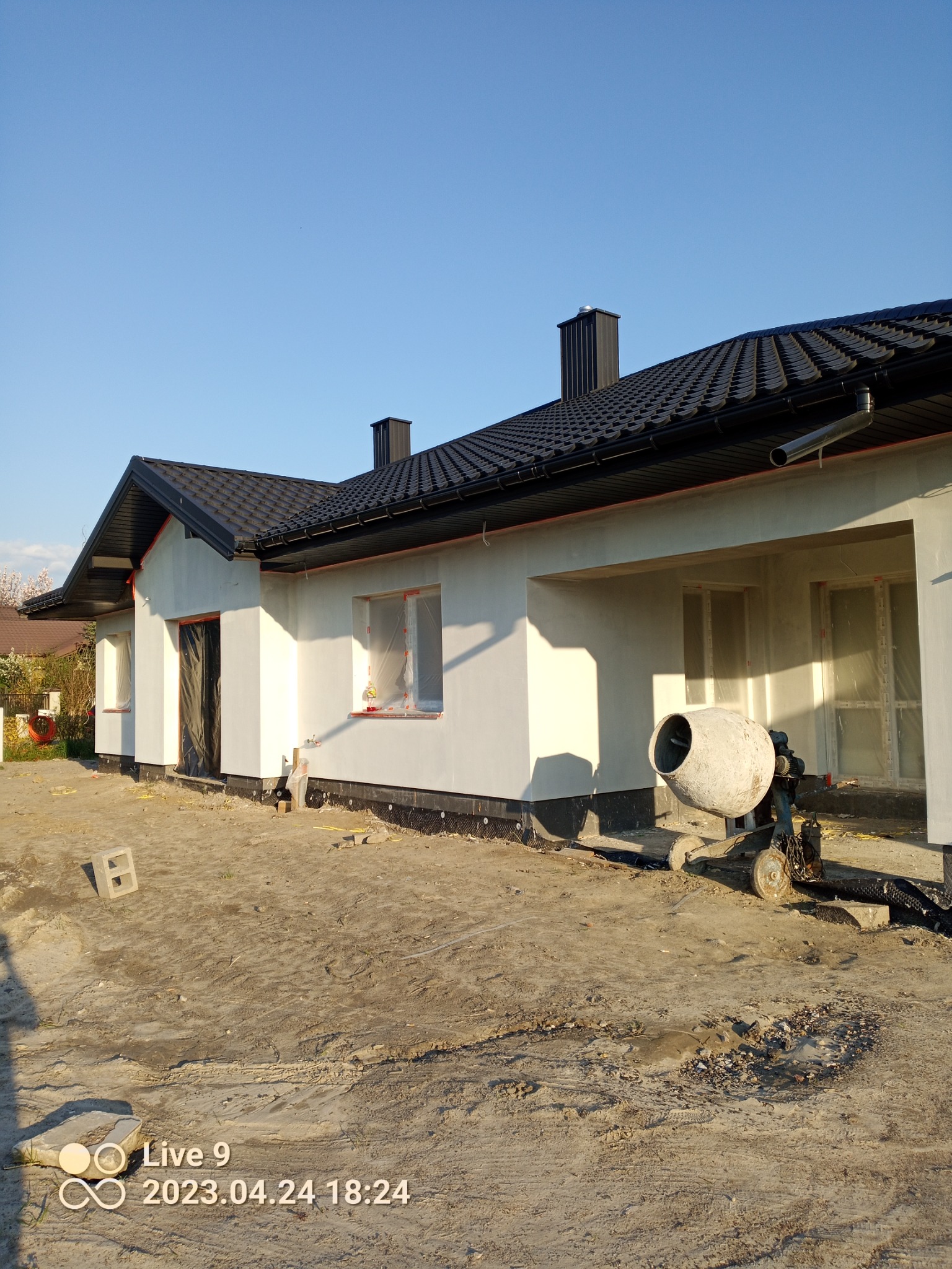 Dom jednorodzinny w trakcie prac elewacyjnych, widoczna świeżo położona warstwa tynku, zabezpieczone okna folią, obok stoi betoniarka, teren wokół domu przygotowany pod dalsze prace.