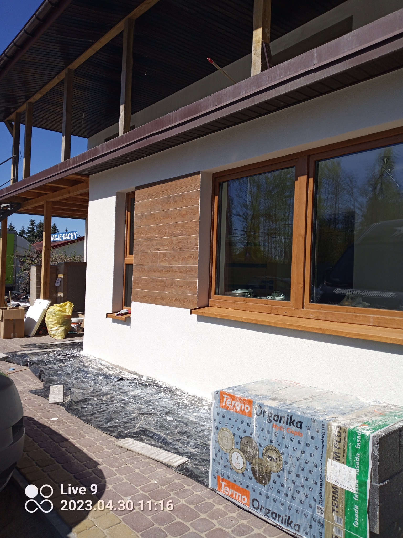 Wykończona elewacja domu z oknami w drewnianej ramie i dekoracyjnym panelem imitującym drewno, obok paleta z materiałem izolacyjnym Termo Organika, teren zabezpieczony folią budowlaną.