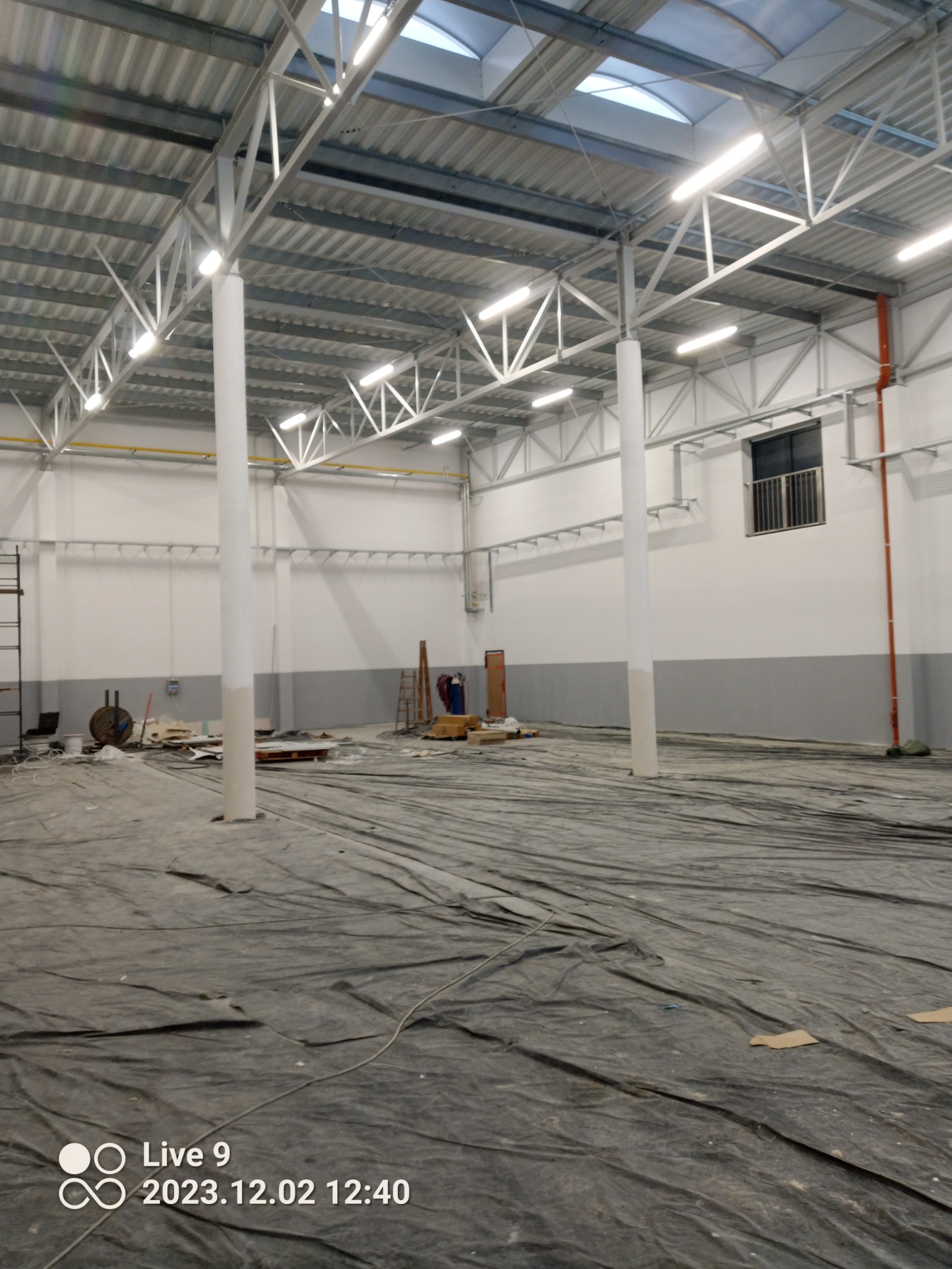 Wnętrze hali przemysłowej w trakcie prac wykończeniowych, podłoga zabezpieczona folią malarską, widoczne białe filary, sufit z metalową konstrukcją i oknami dachowymi, ściany częściowo pomalowane...