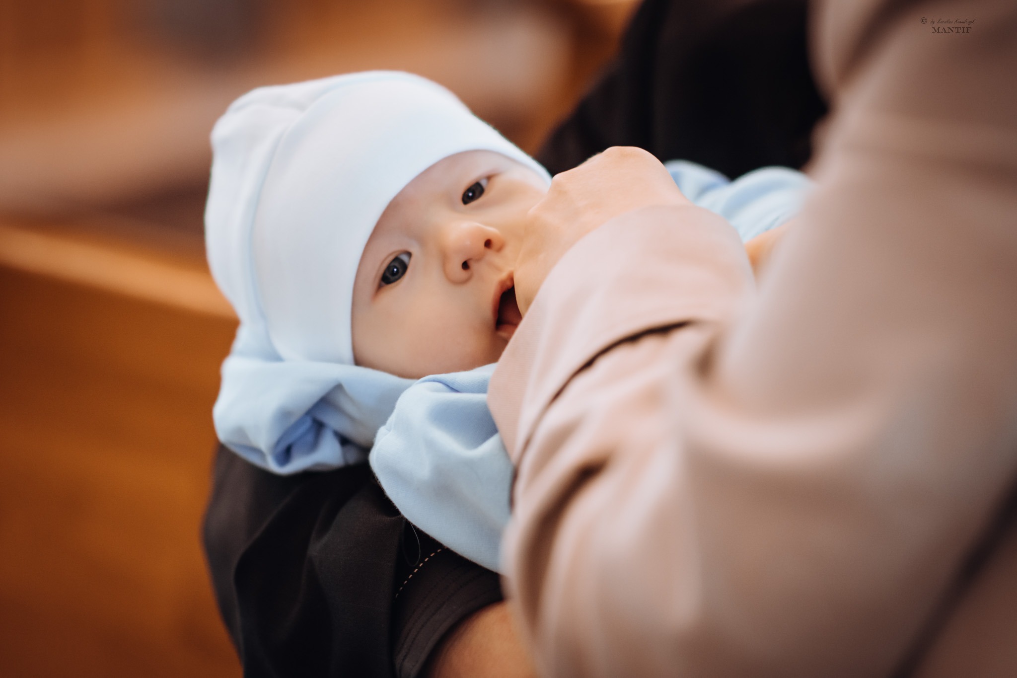 Niemowlę w białym czepku i jasnoniebieskim ubranku, trzymane przez osobę w beżowej odzieży, z otwartymi ustami i zaciekawionym spojrzeniem.
