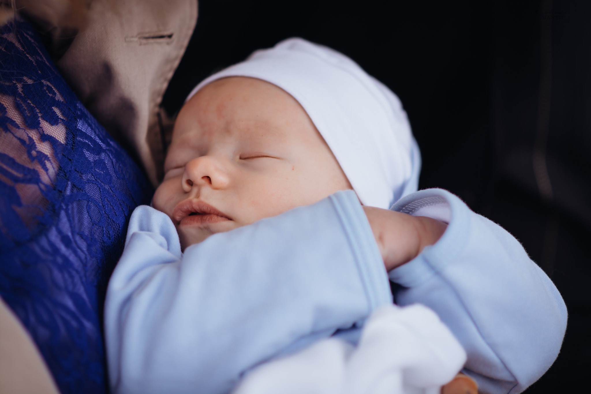 Śpiący noworodek w jasnoniebieskim ubranku i białej czapeczce, trzymany na rękach osoby ubranej w kobaltową koronkową bluzkę i beżowy płaszcz.