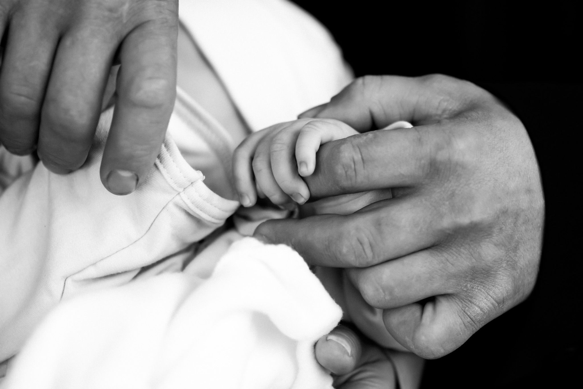 Czarno-białe zbliżenie dłoni noworodka trzymanej przez dłoń dorosłego na tle białego ubranka.