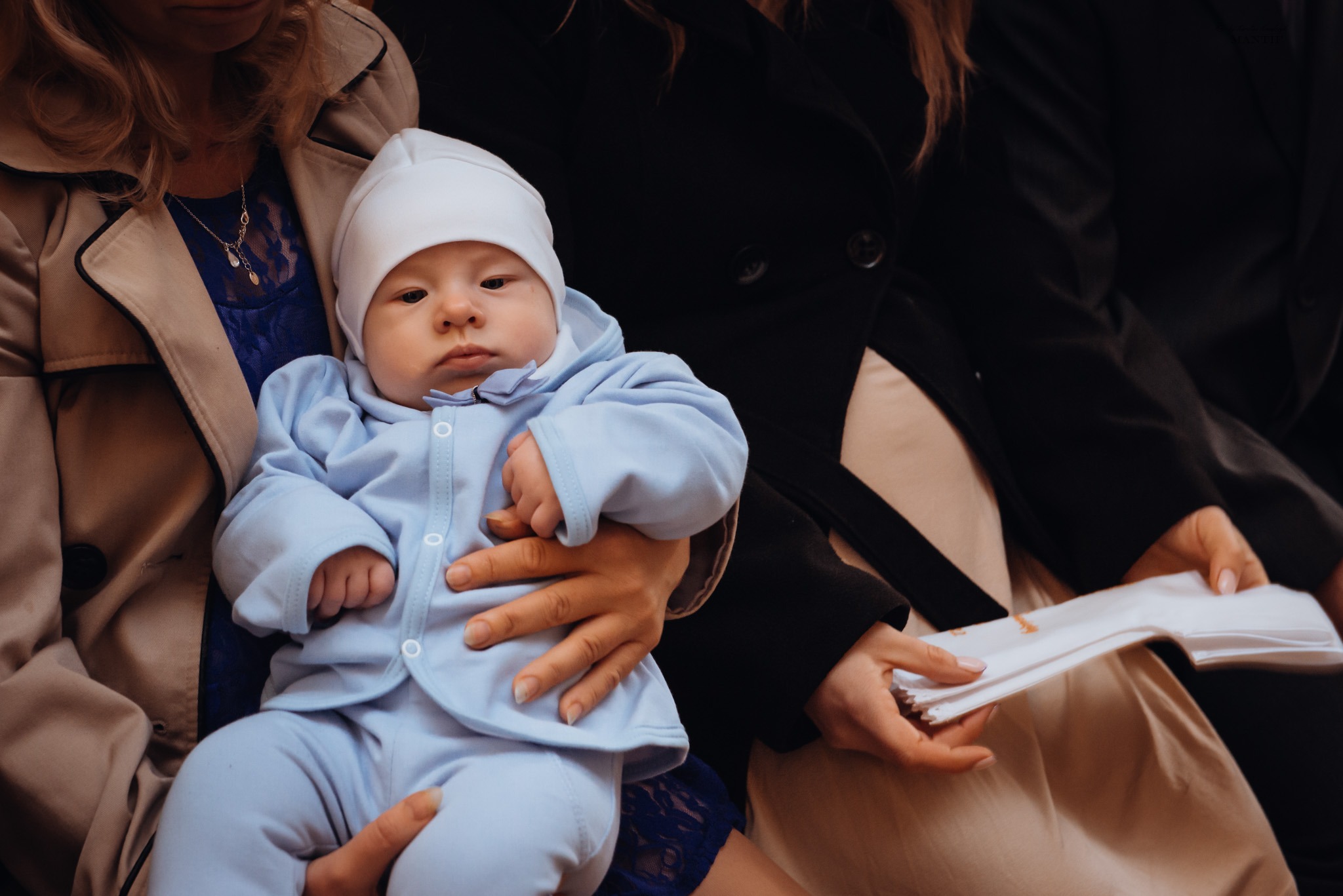 Niemowlę w błękitnym ubranku i białej czapeczce, trzymane na rękach przez kobietę z delikatnym naszyjnikiem, w tle osoby w ciemnych płaszczach.