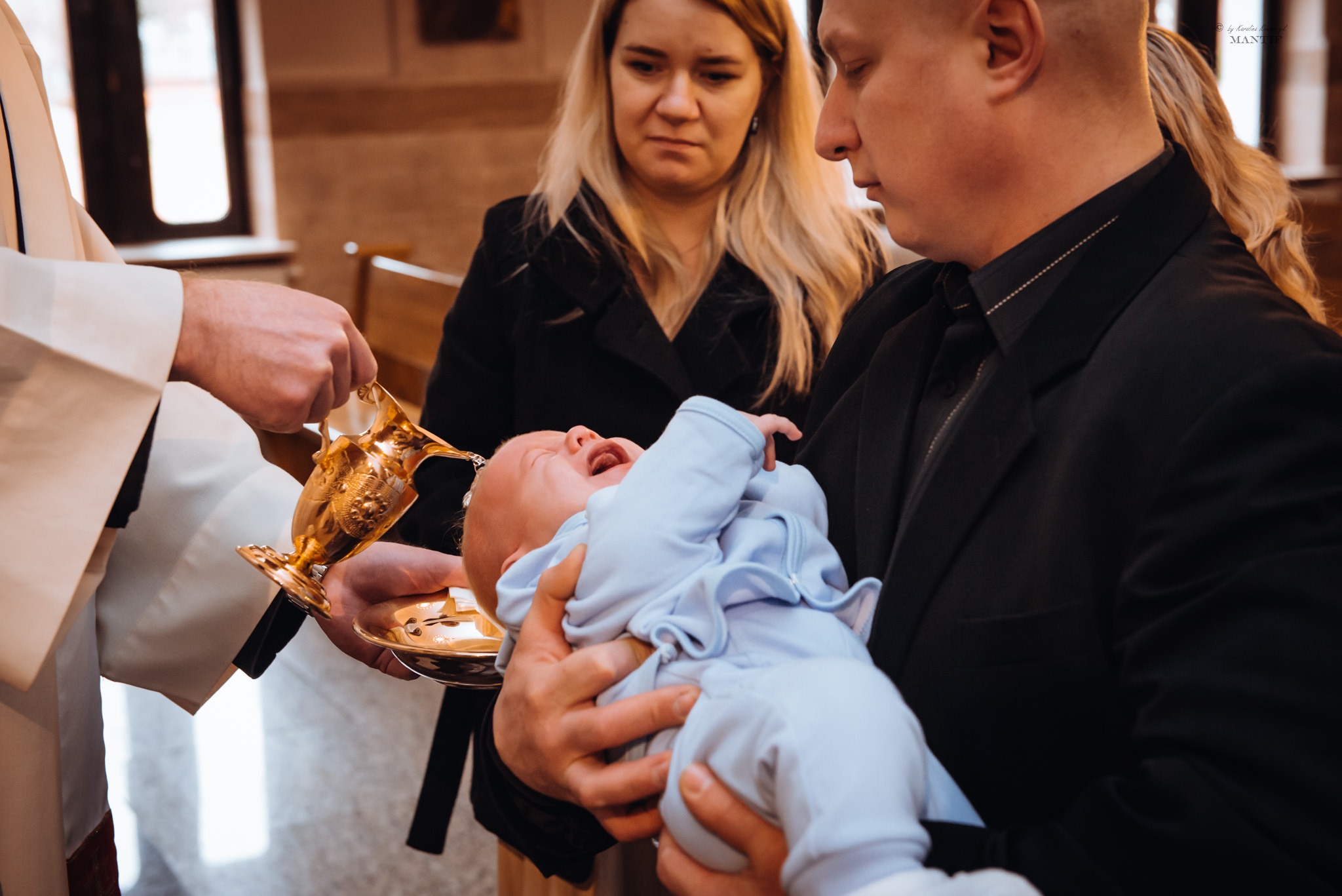 Ksiądz polewa wodą głowę płaczącego niemowlęcia trzymanego przez ojca podczas ceremonii w kościele, w tle stoi matka dziecka.