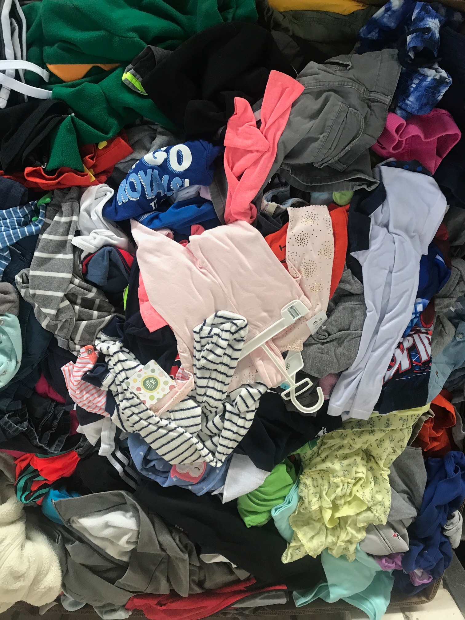 Stos różnorodnych ubrań, w tym koszulki z nadrukiem, spodnie cargo, bluzki w paski, sukienki z falbanami, niektóre na wieszakach, tworzące chaotyczny, kolorowy wzór.