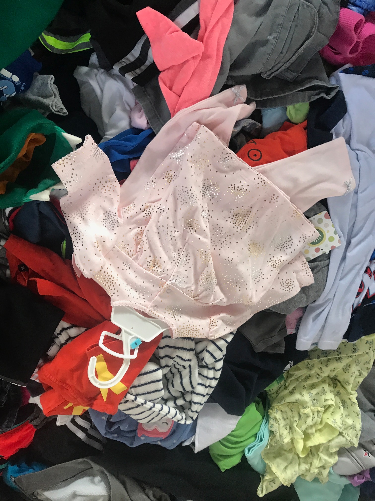 Stos różnorodnych ubrań, w tym bluzka dziecięca w kolorze jasnoróżowym z motywem złotych gwiazdek, leżąca na wierzchu innych elementów garderoby, widoczny fragment wieszaka.