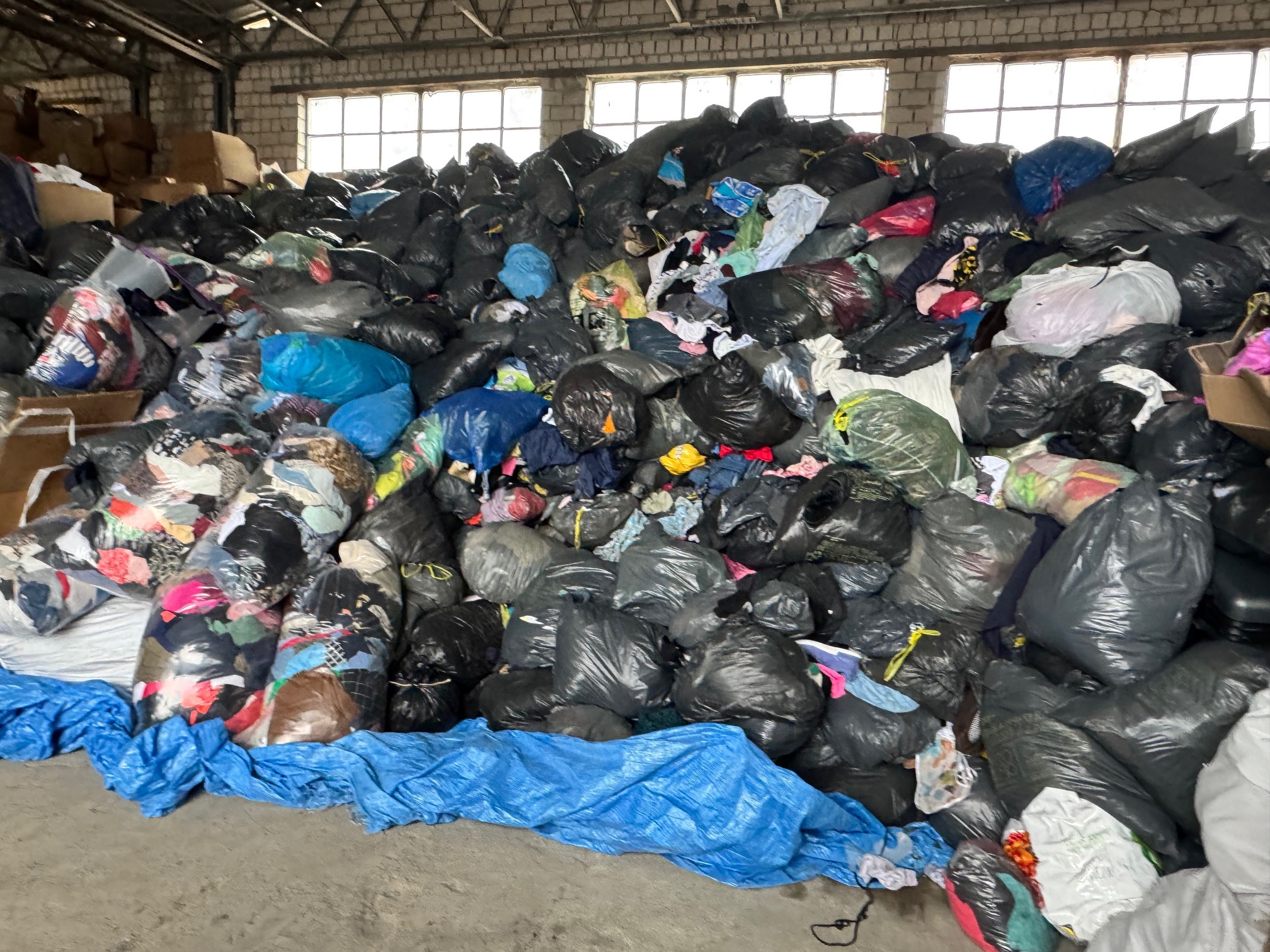 Sterta czarnych worków z odzieżą w magazynie z ceglanymi ścianami i oknami w tle, przykryta częściowo niebieską folią.