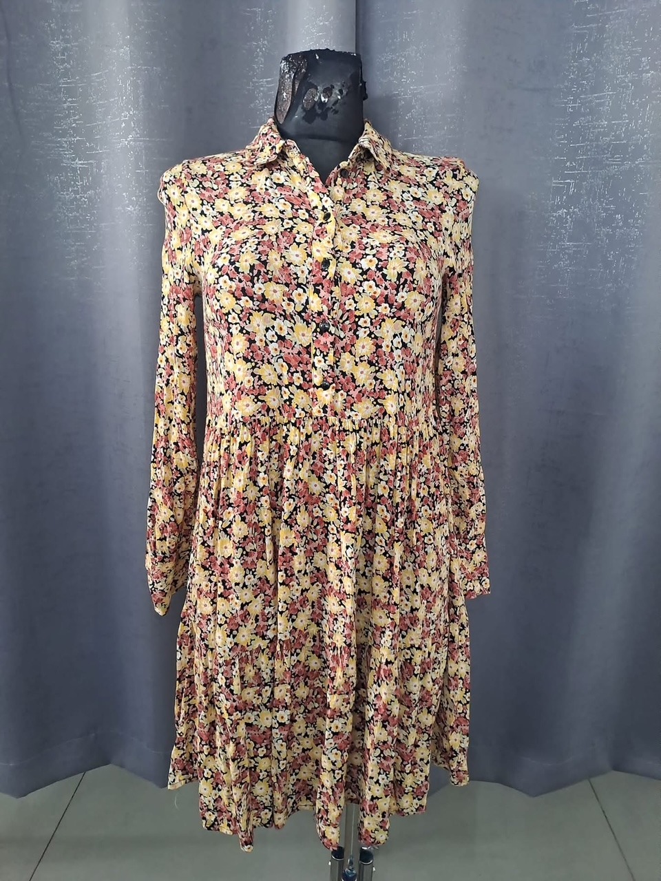 Sukienka damska w drobne kwiaty na manekinie, z długim rękawem i kołnierzykiem, na tle szarej zasłony.