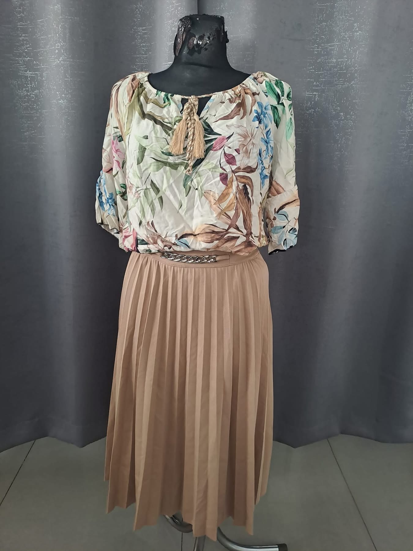 Manekin prezentuje stylizację: bluzka w kwiaty z ozdobnymi sznurkami i beżowa plisowana spódnica z łańcuszkowym paskiem.