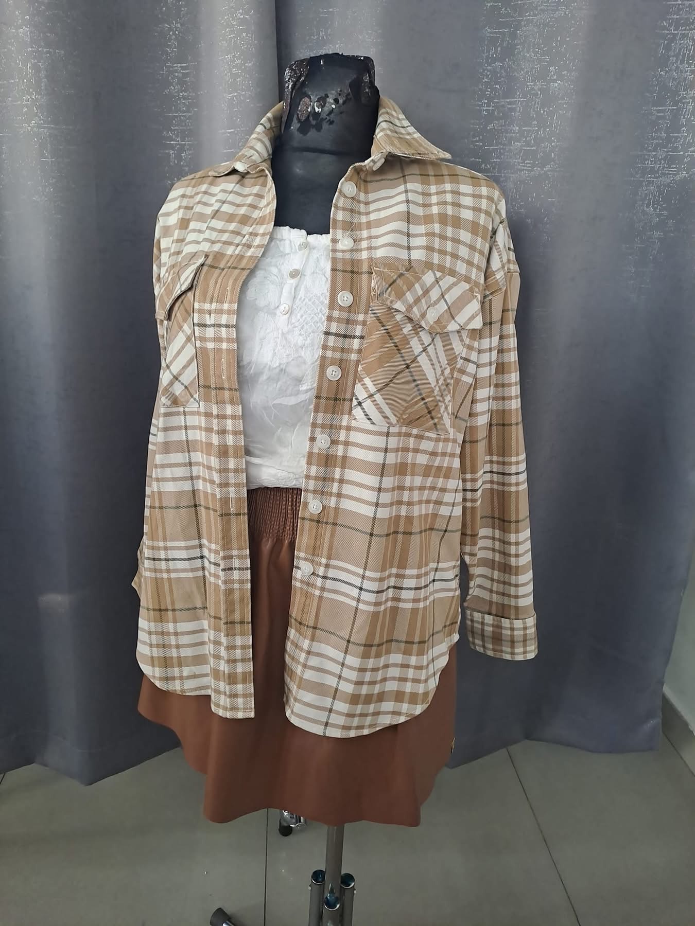 Manekin prezentuje stylizację: rozpiętą koszulę w beżowo-białą kratę z kieszeniami, białą bluzkę z koronkowymi detalami i brązową spódnicę z imitacji skóry.