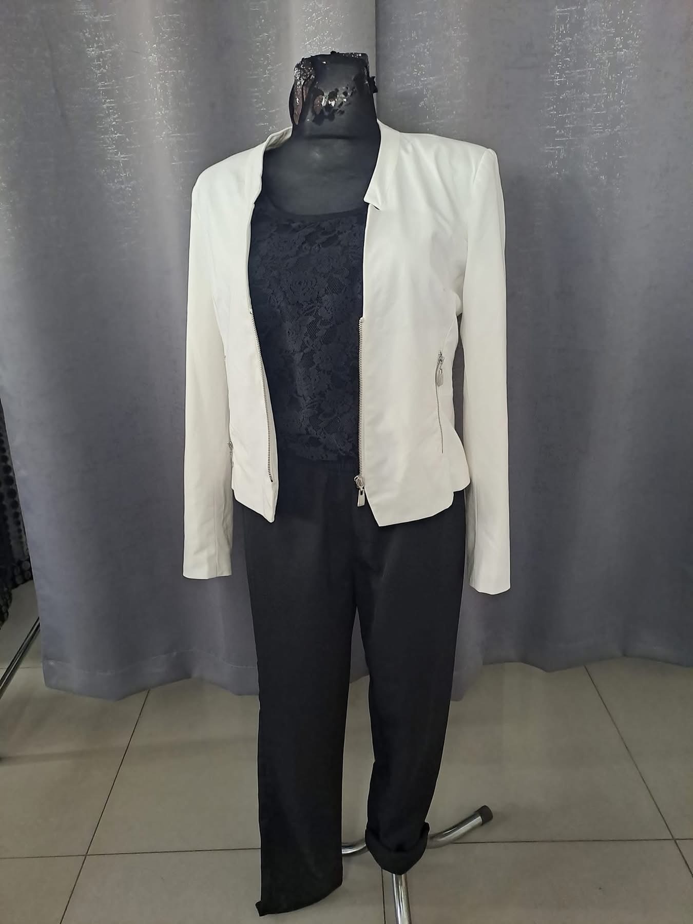 Manekin prezentuje zestaw: biała kurtka z zamkiem, czarna bluzka z koronką i czarne spodnie. Tło stanowi szara zasłona.