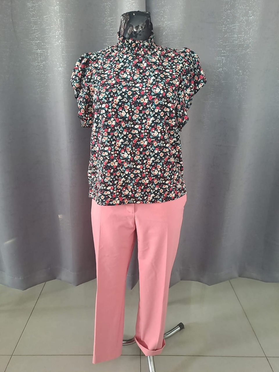 Manekin prezentuje bluzkę w drobne kwiaty z golfem i krótkim rękawem oraz różowe spodnie z podwiniętymi nogawkami na tle szarej zasłony.