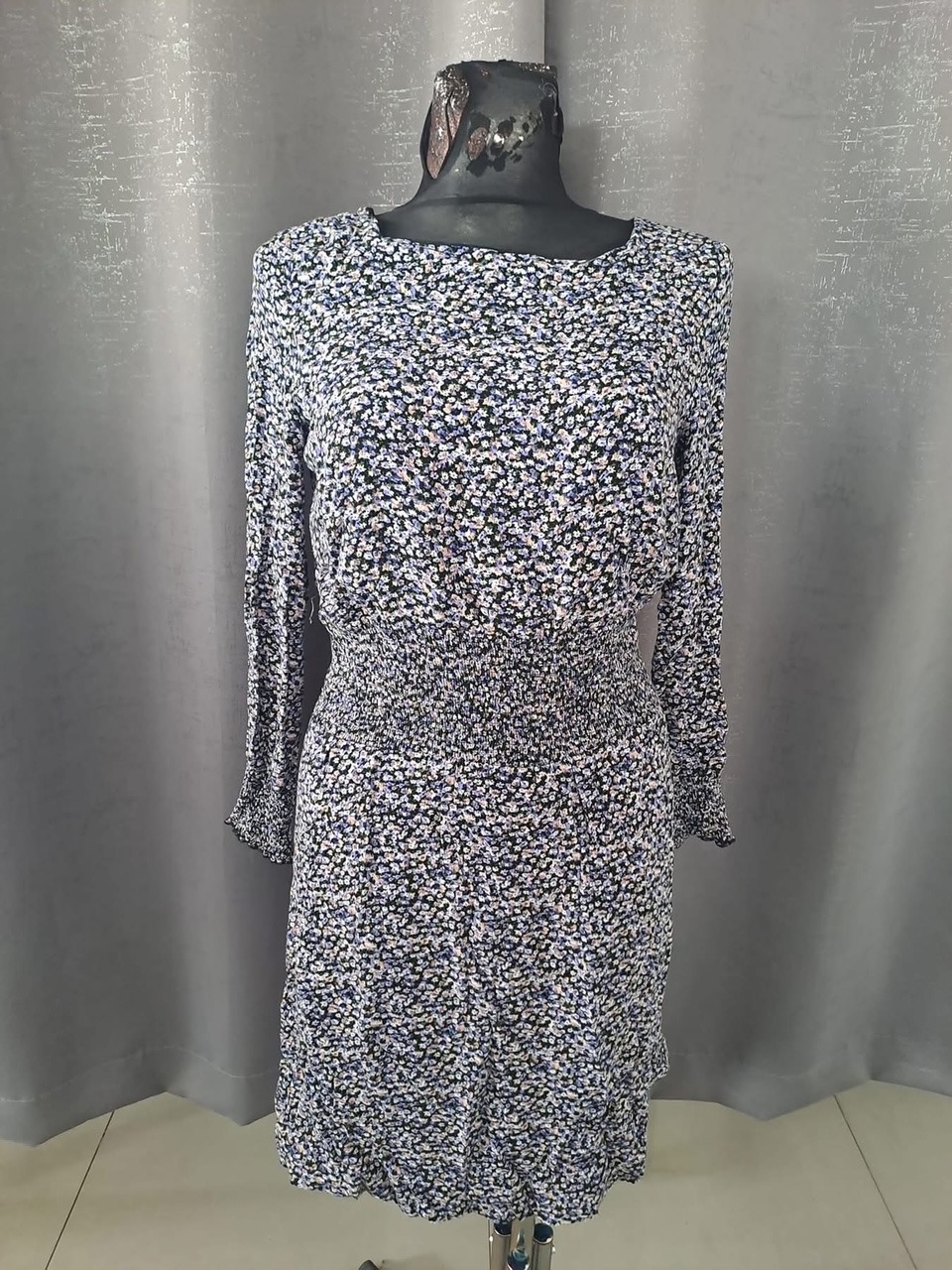 Krótka sukienka w drobne kwiatki na manekinie z szarym tłem. Sukienka ma długie rękawy i elastyczny pas.