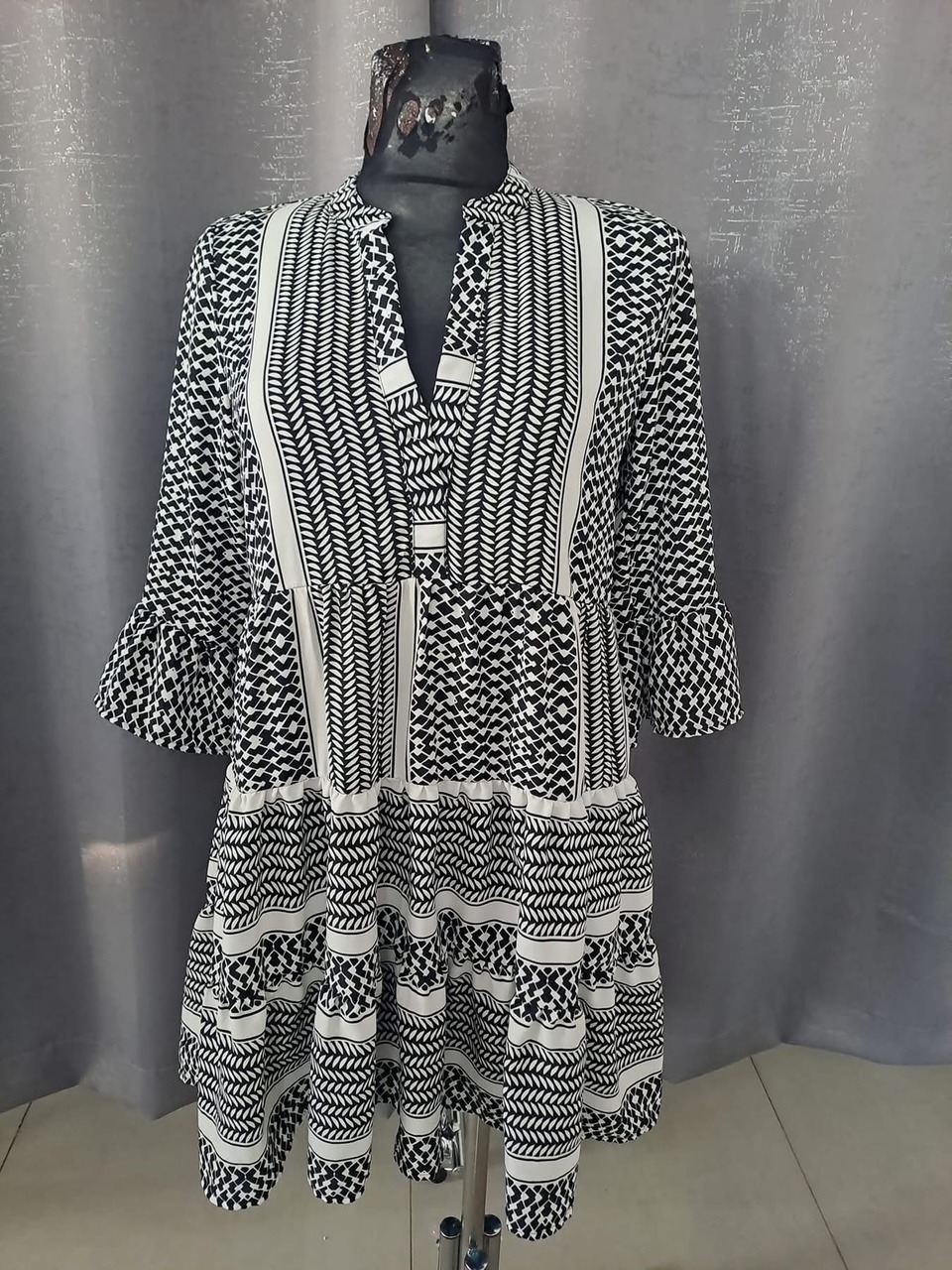 Czarno-biała sukienka z wzorem geometrycznym na manekinie, z falbankami na dole i rękawach, na tle szarej zasłony.