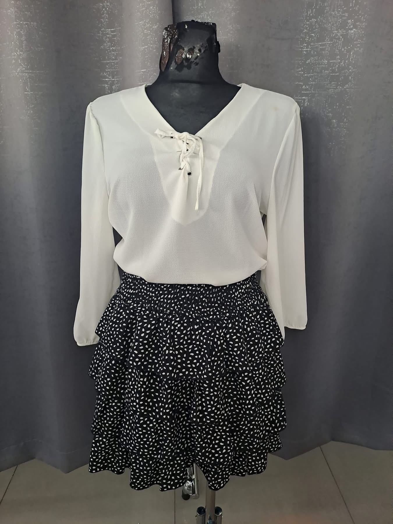 Manekin prezentuje białą bluzkę z długim rękawem i sznurkowanym dekoltem oraz czarną spódnicę w białe wzory z falbanami.