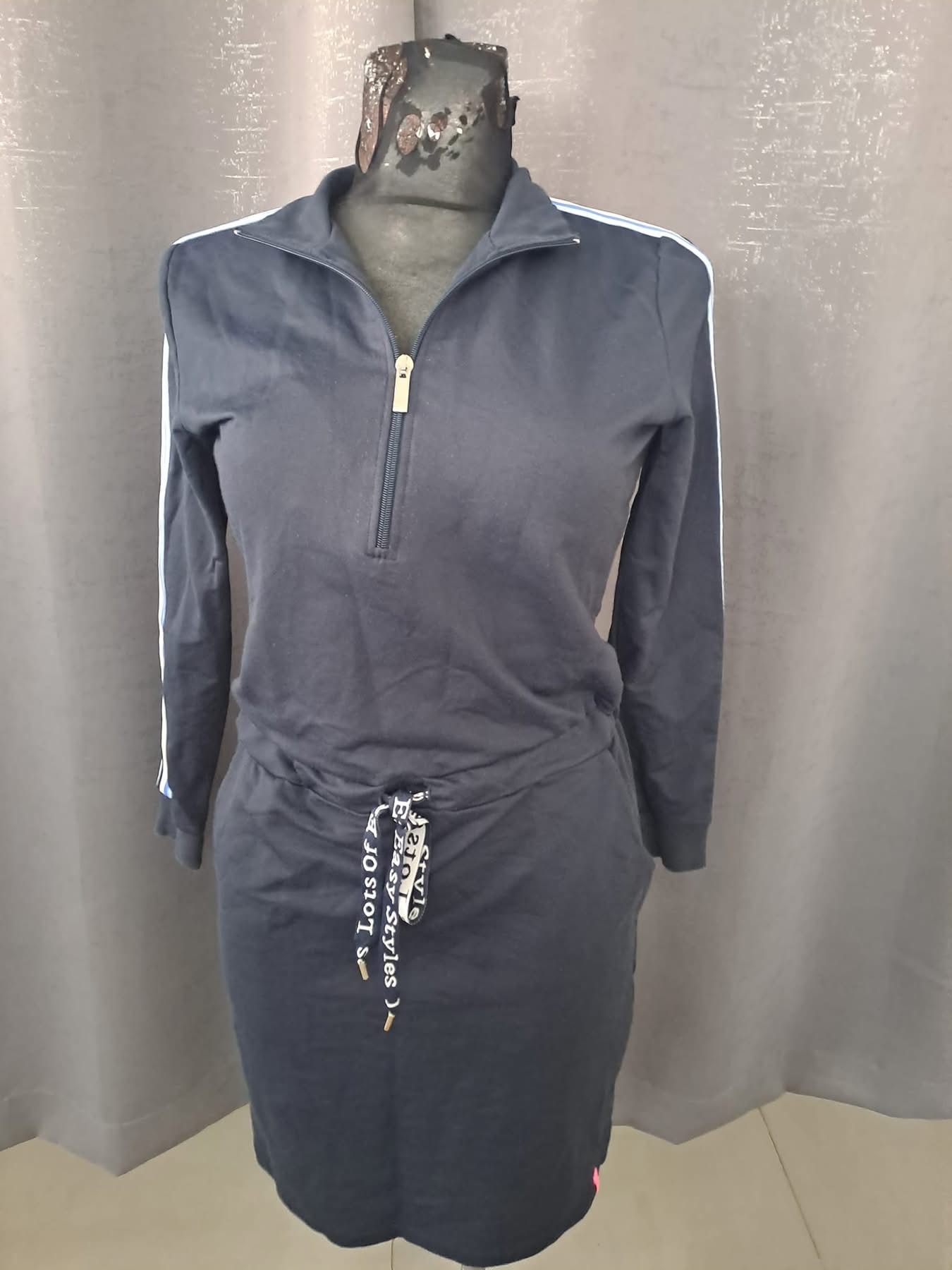 Granatowa sukienka dresowa z białymi lampasami na rękawach, zapinana na zamek, z ozdobnym sznurkiem w pasie, prezentowana na manekinie na tle szarej zasłony.