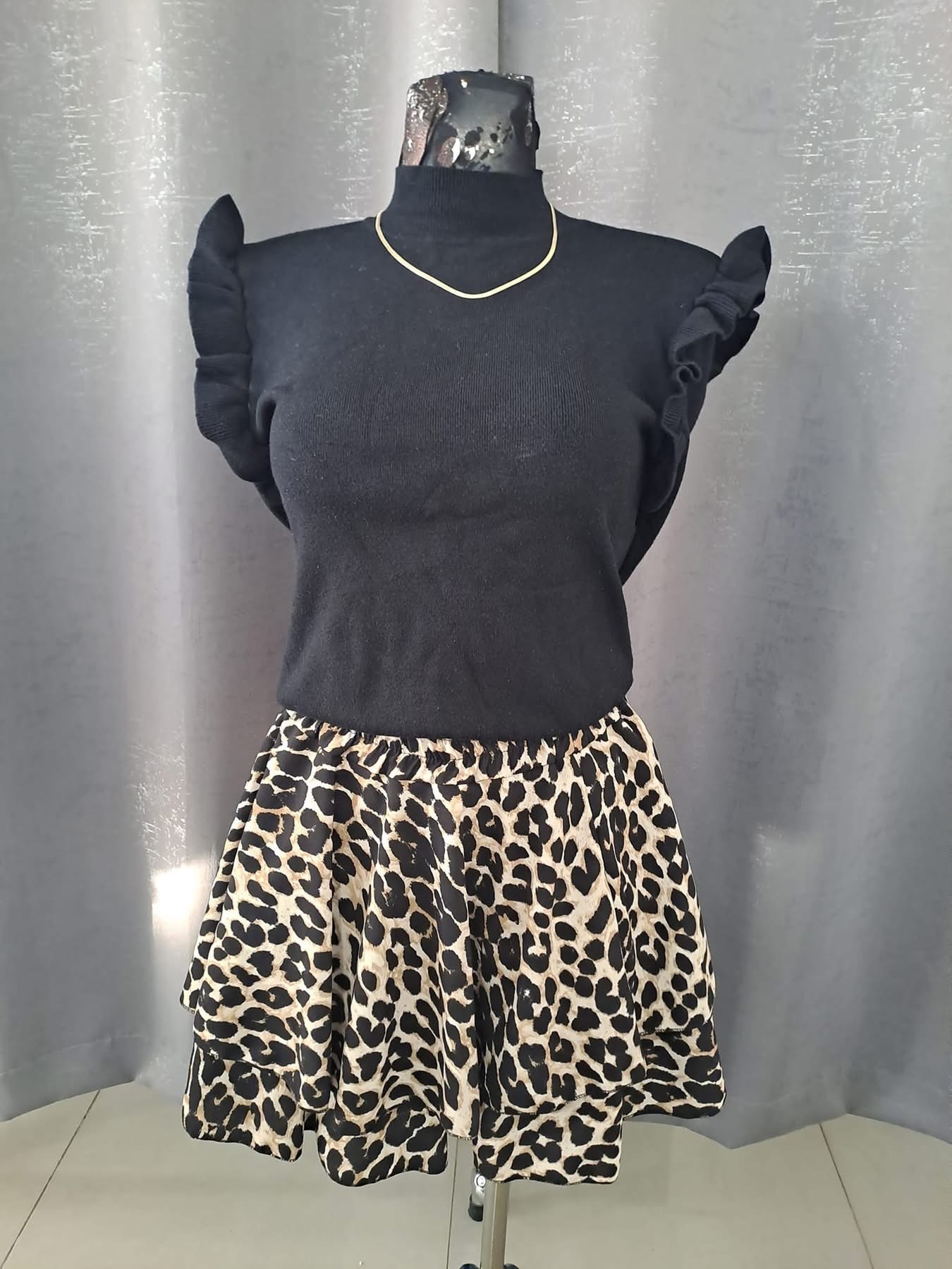 Czarny golf z falbankami na ramionach i spódnica w panterkę na manekinie, złoty łańcuszek na szyi, szare tło.