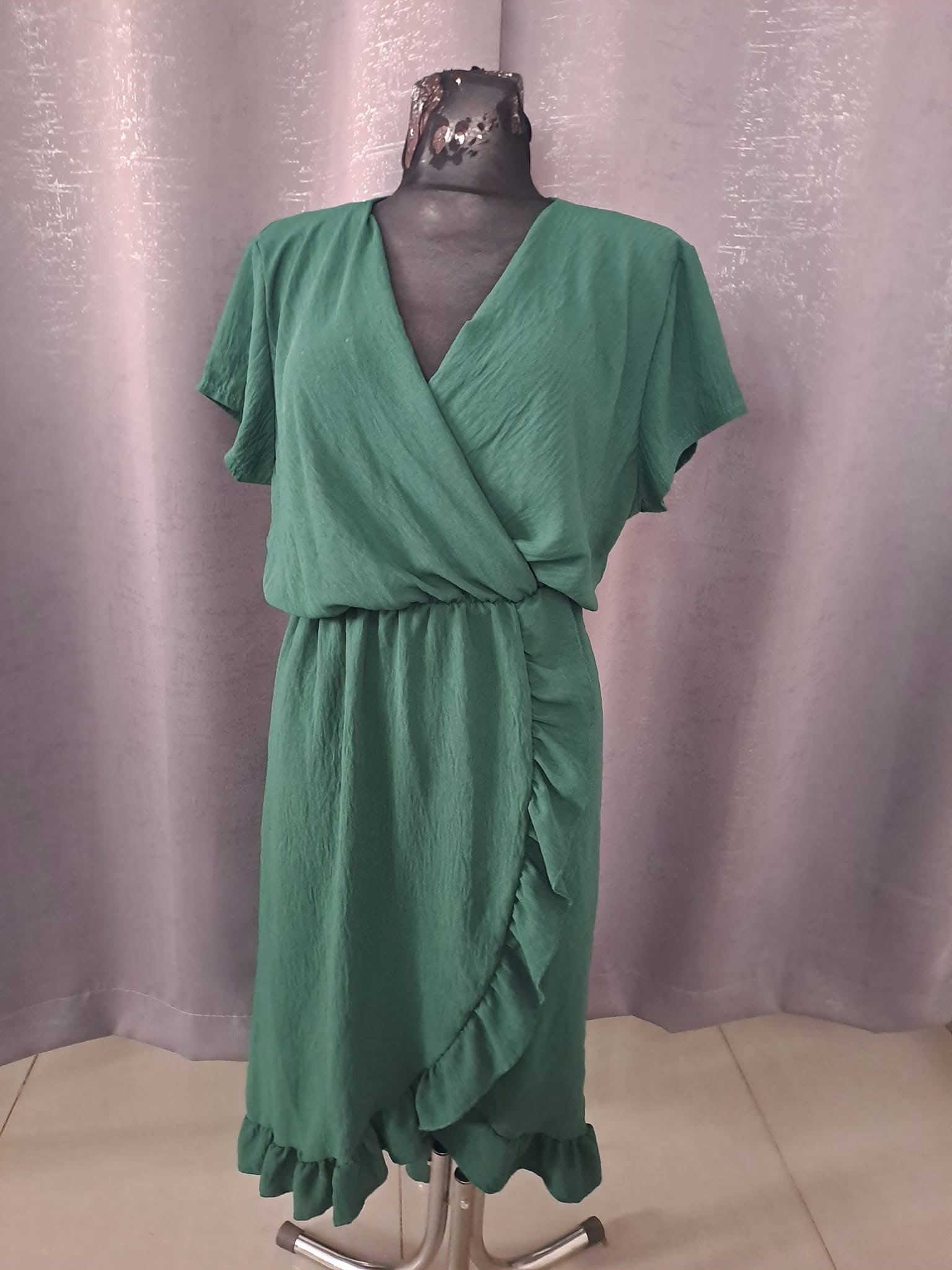 Zielona sukienka z krótkim rękawem, drapowana w talii i z falbaną, prezentowana na manekinie na tle szarej zasłony.