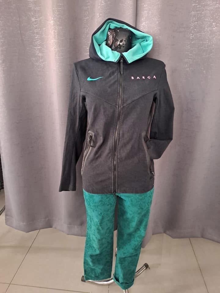 Szary komplet dresowy z kapturem i zielonym wykończeniem, z logo i napisem 'Barca', prezentowany na manekinie na tle szarej zasłony.