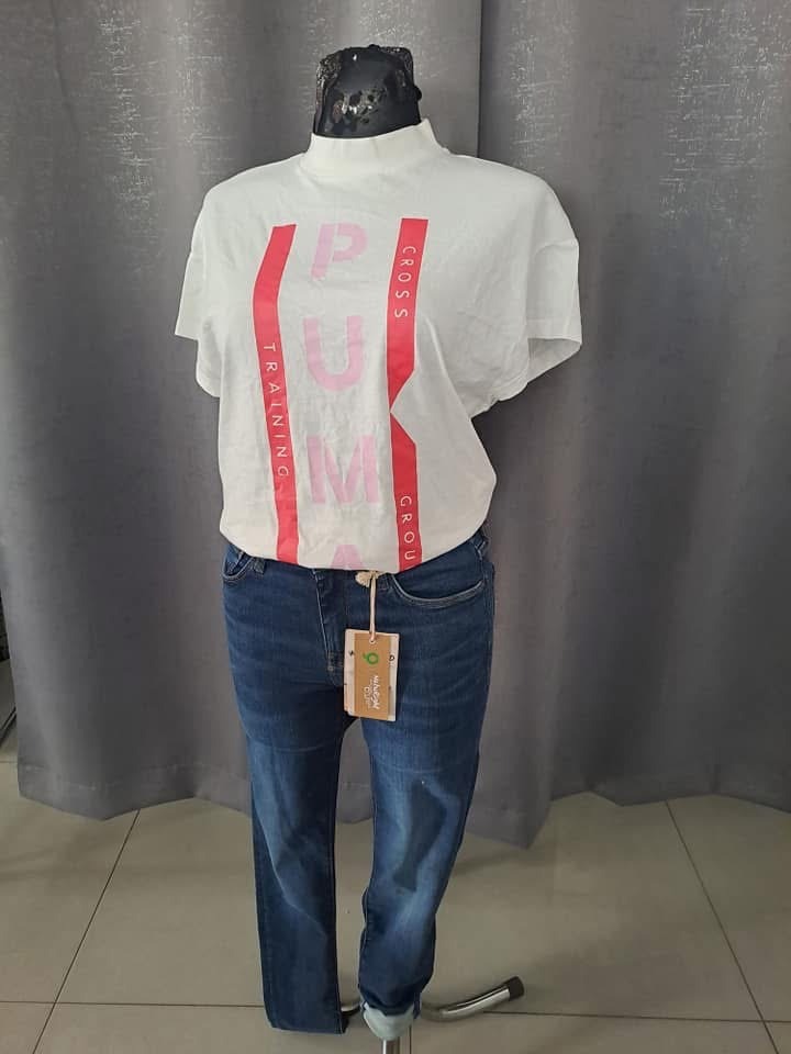 Manekin prezentuje białą bluzkę z napisem 'Puma Cross Training Group' i niebieskie jeansy z metką, na tle szarej zasłony.