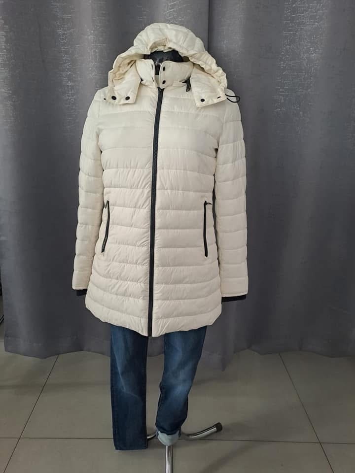 Kremowa, pikowana kurtka damska z kapturem, zapinana na czarny zamek błyskawiczny, prezentowana na manekinie z jeansami.