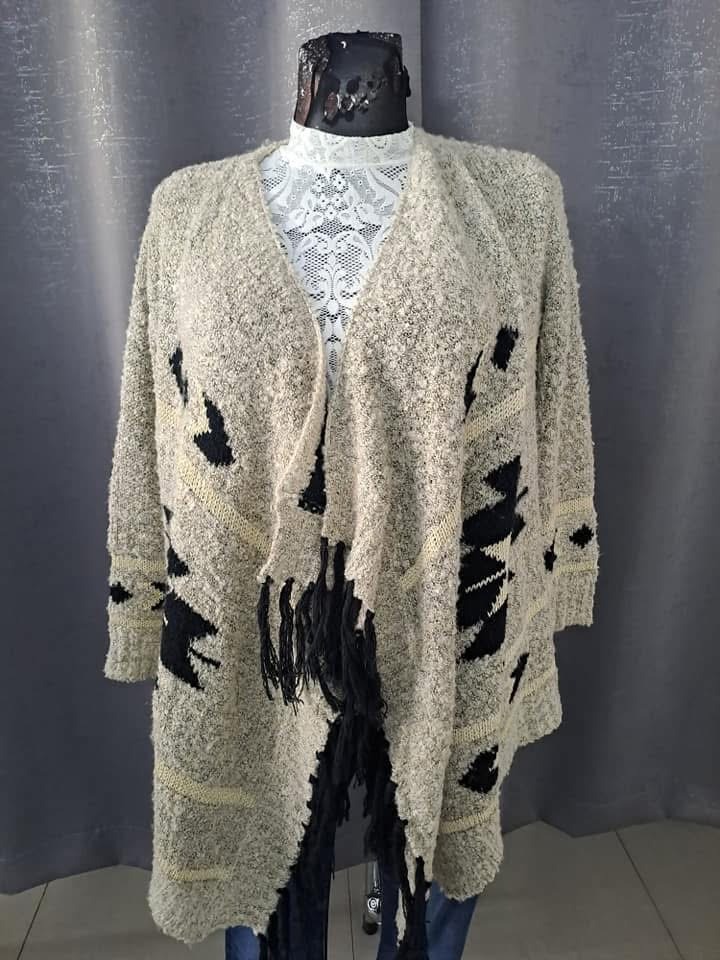 Beżowy, dziergany kardigan z czarnymi, geometrycznymi wzorami i frędzlami, prezentowany na manekinie z koronkową bluzką, na tle szarej zasłony.
