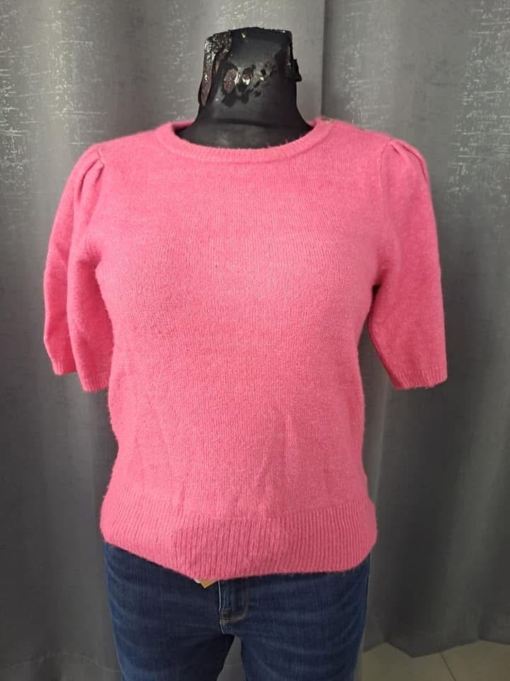 Różowy sweter z krótkim rękawem i bufkami na ramionach, prezentowany na manekinie z dżinsami, na tle szarej zasłony.