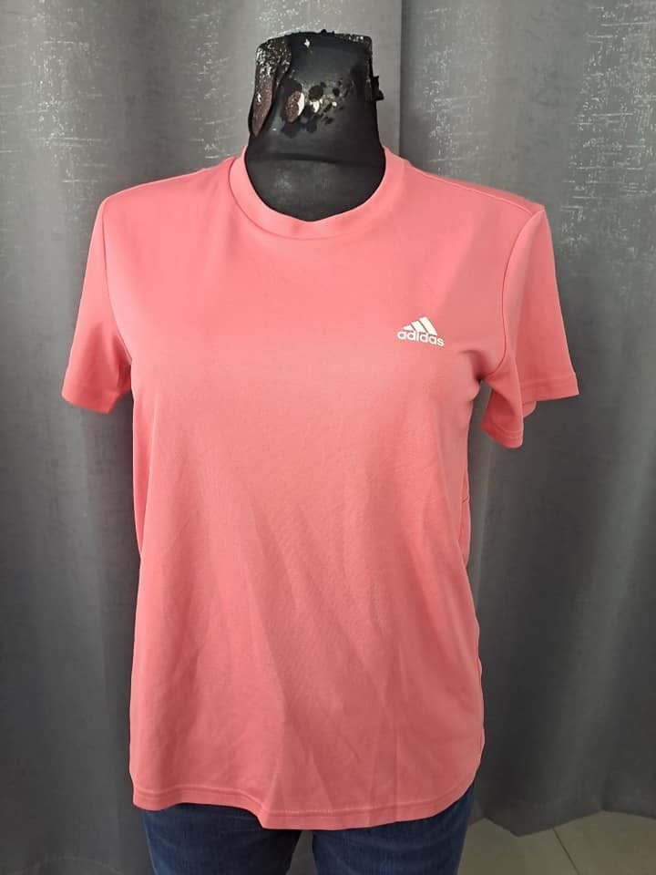 Koralowa koszulka sportowa Adidas na manekinie z widocznym logo, tło stanowi szara zasłona.