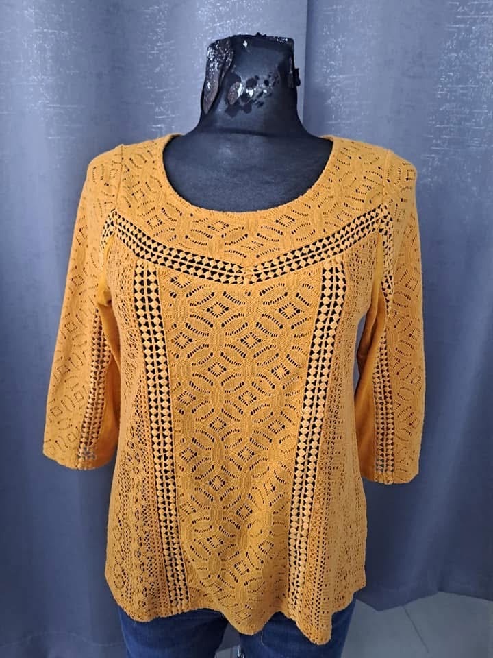 Musztardowa, ażurowa bluzka z rękawami 3/4 na manekinie, z geometrycznym wzorem i okrągłym dekoltem.