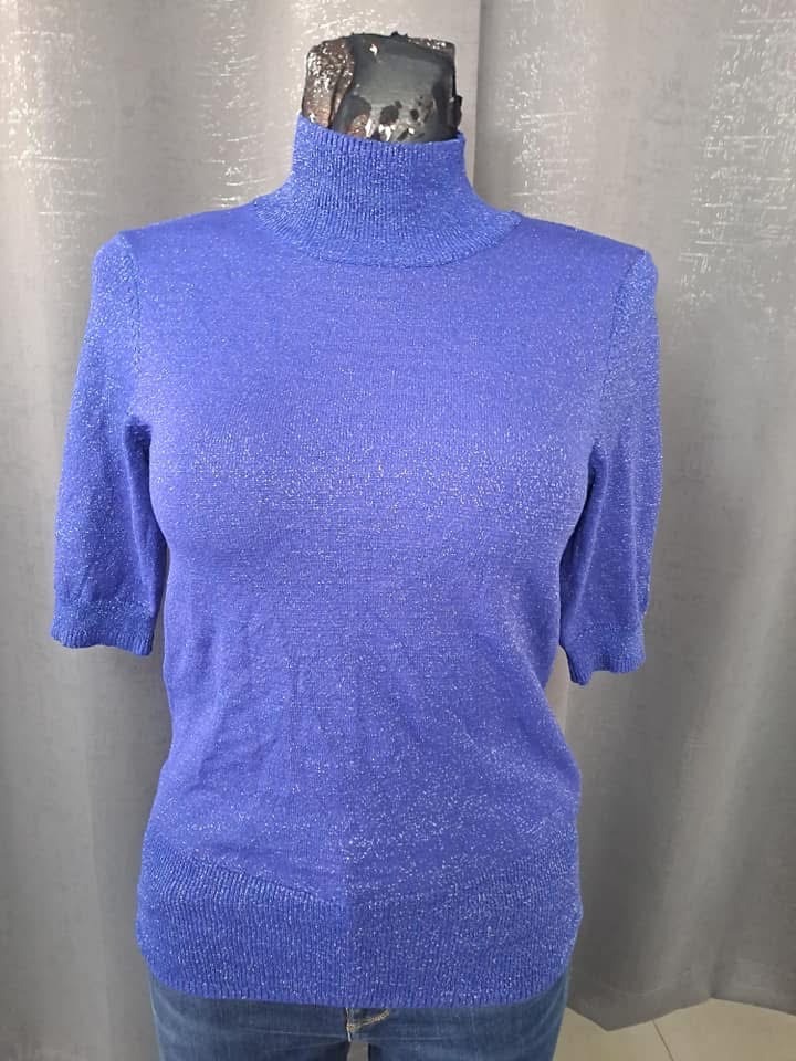 Błękitny sweter z krótkim rękawem i golfem, z delikatnym połyskiem, prezentowany na manekinie, na tle szarej zasłony.