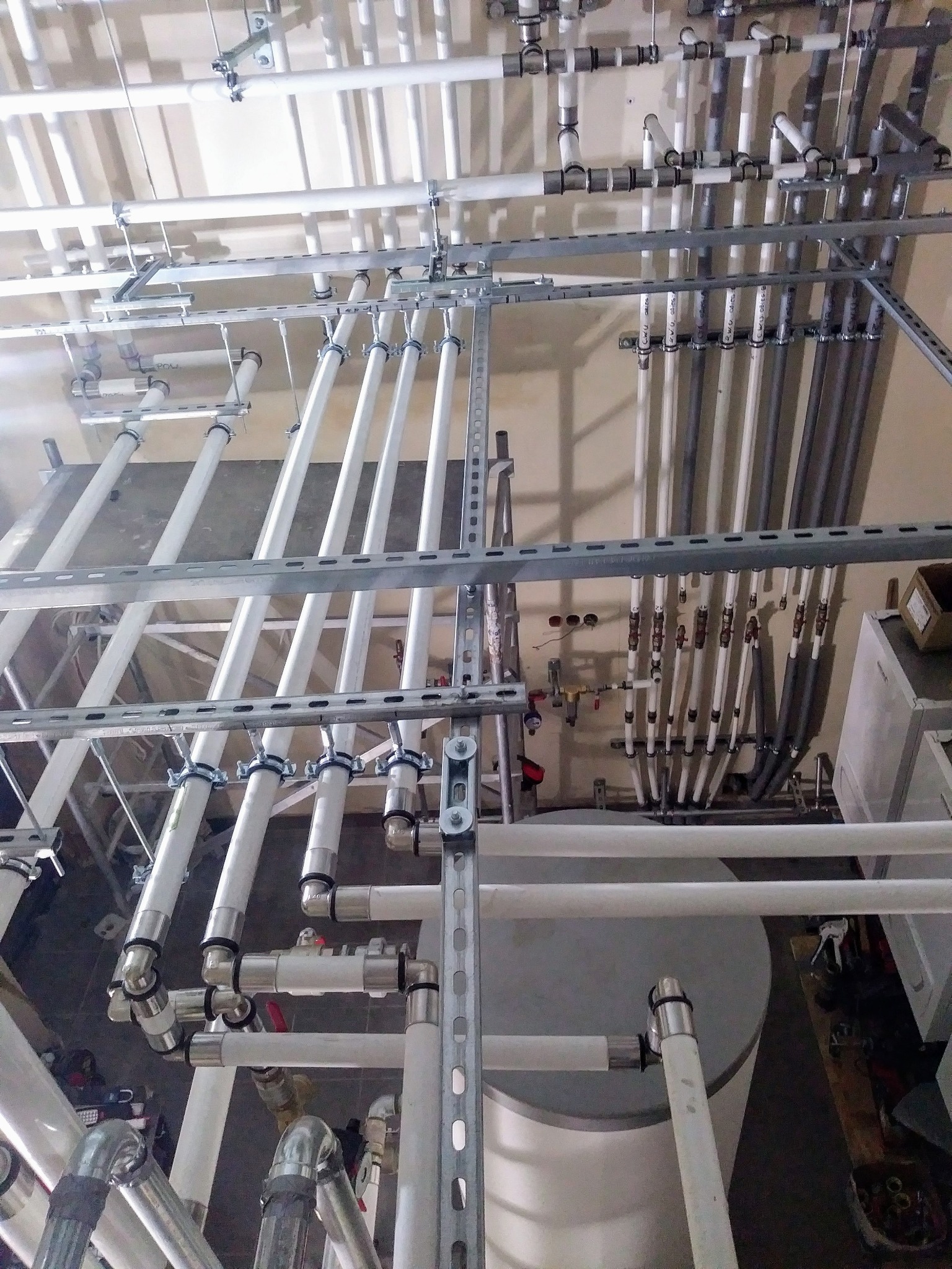 Skomplikowany system rur instalacji hydraulicznej i grzewczej pod sufitem, z widocznymi elementami mocującymi i drabinką w tle, w pomieszczeniu technicznym.