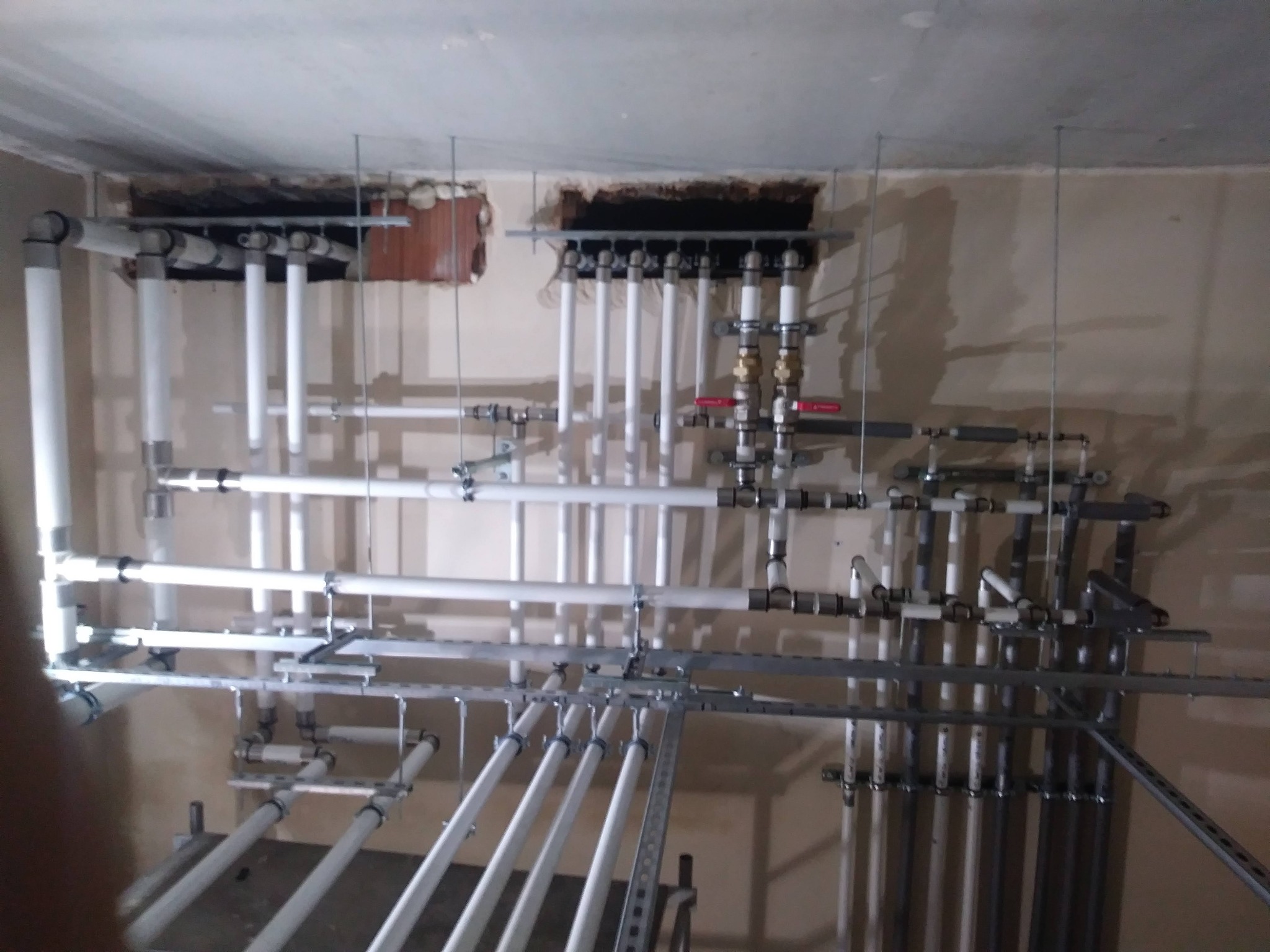 Rozbudowana instalacja rur w budynku w trakcie montażu, widoczne białe i szare rury, metalowe wsporniki, otwory w suficie z instalacjami.