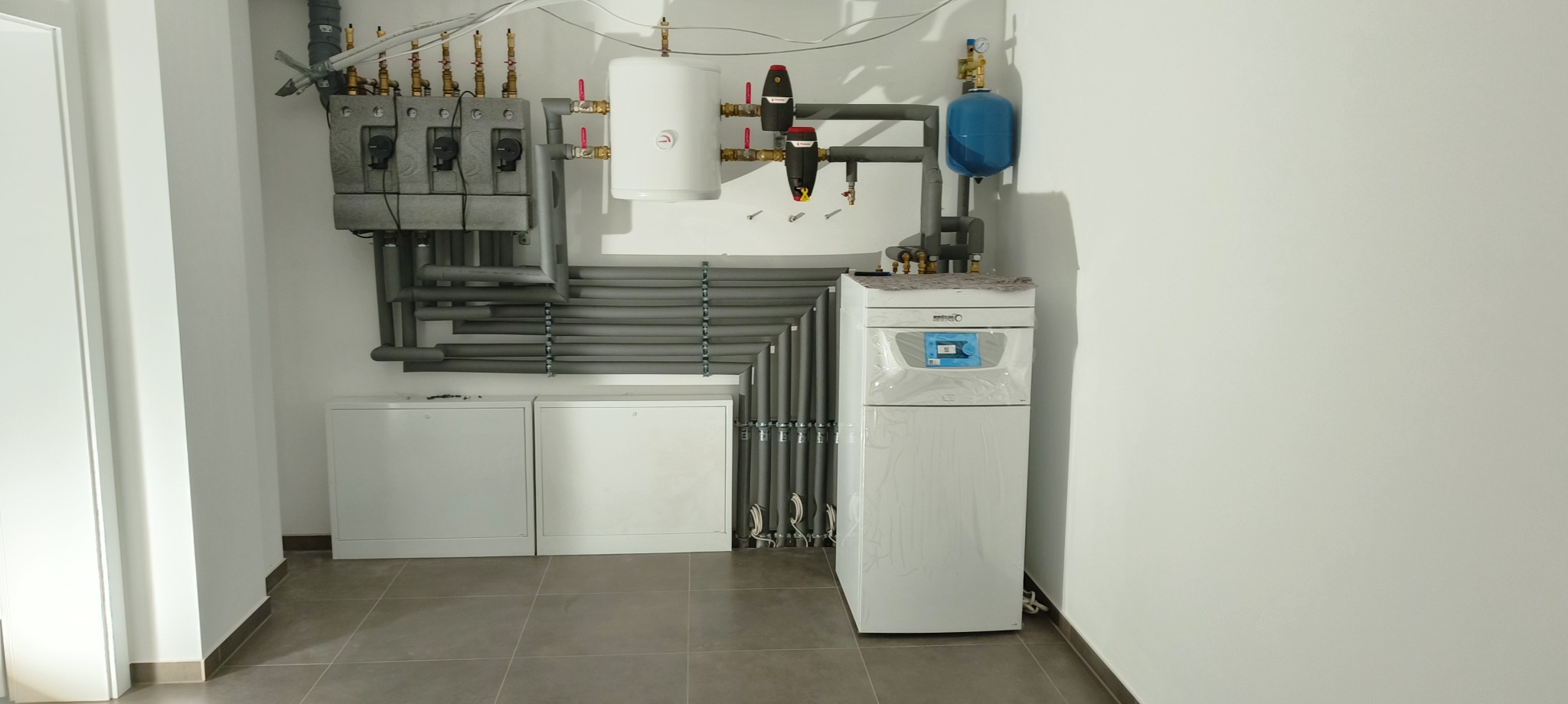 Nowoczesna kotłownia w jasnym pomieszczeniu z szarymi rurami w izolacji, białym piecem gazowym w folii ochronnej oraz zbiornikami wyrównawczymi i rozdzielaczami na ścianie.