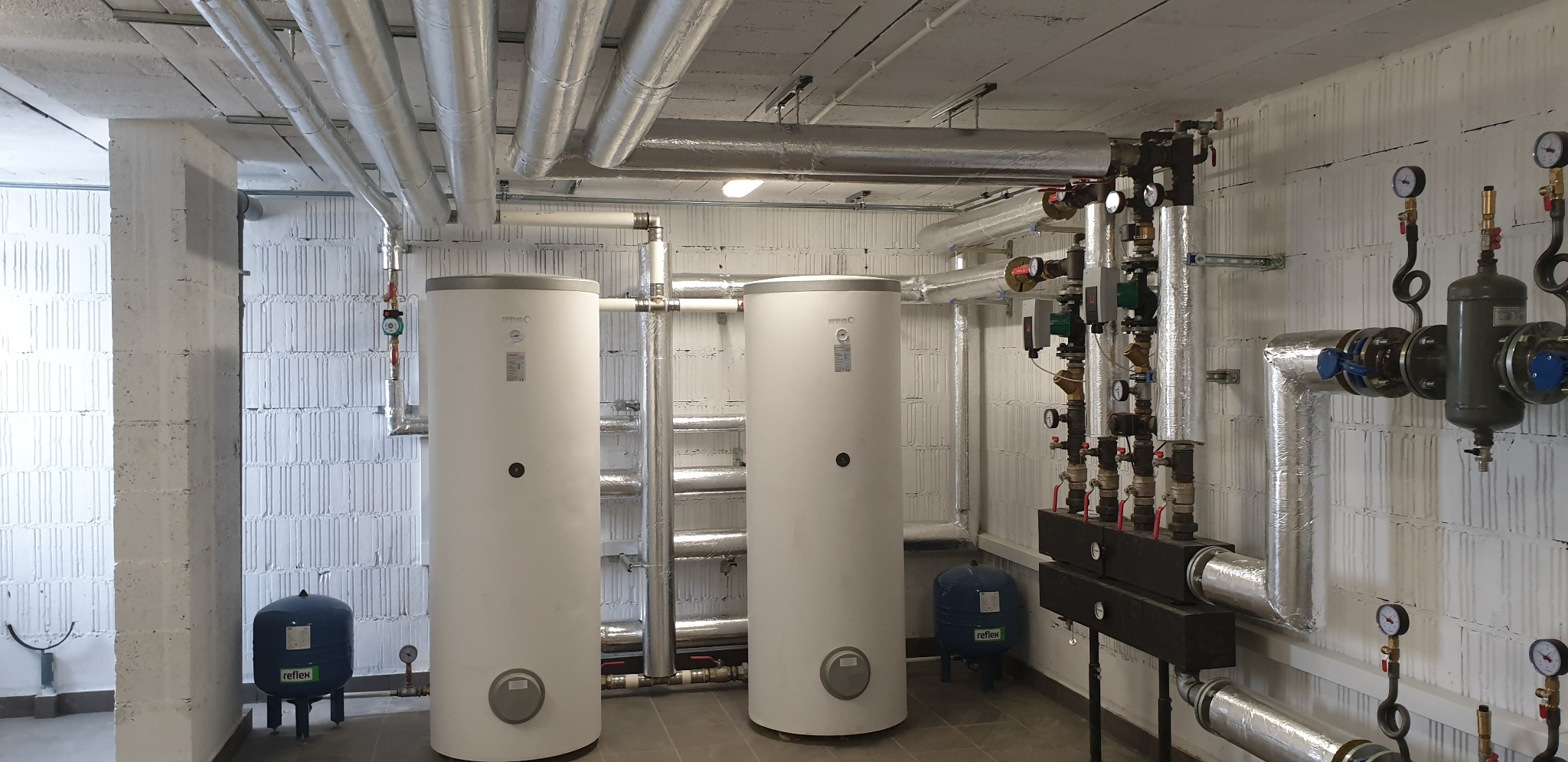 Dwa białe zbiorniki buforowe Reflex w kotłowni, z widocznymi rurami w izolacji termicznej, manometrami i elementami instalacji grzewczej na tle ściany z bloczków betonowych.
