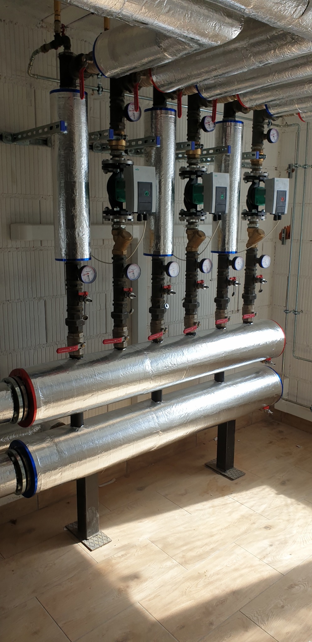 Rozdzielacz instalacji grzewczej z czterema pompami obiegowymi, rury zaizolowane folią aluminiową, widoczne manometry i zawory, tło stanowi ściana z bloczków betonu komórkowego.