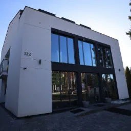 Nowoczesny budynek biurowy z dużymi oknami odbijającymi błękit nieba, numer budynku 122 widoczny na elewacji, otoczony drzewami i brukowanym podjazdem.