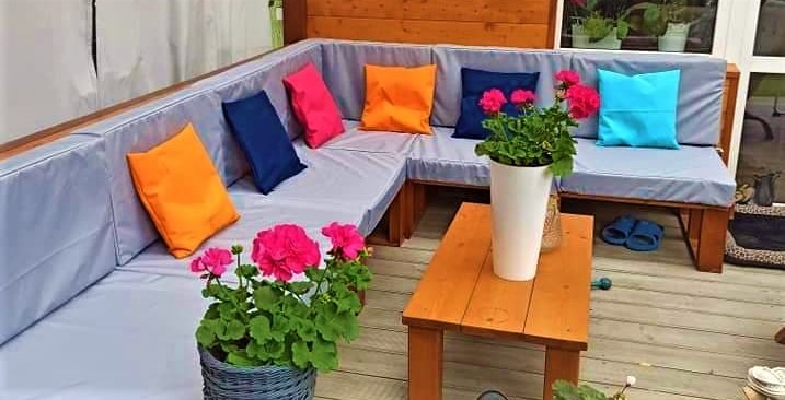 Kącik wypoczynkowy na tarasie z drewnianą sofą w kształcie litery L, jasnoszare poduchy, kolorowe poduszki dekoracyjne (różowa, pomarańczowa, granatowa, błękitna), drewniany stolik kawowy...