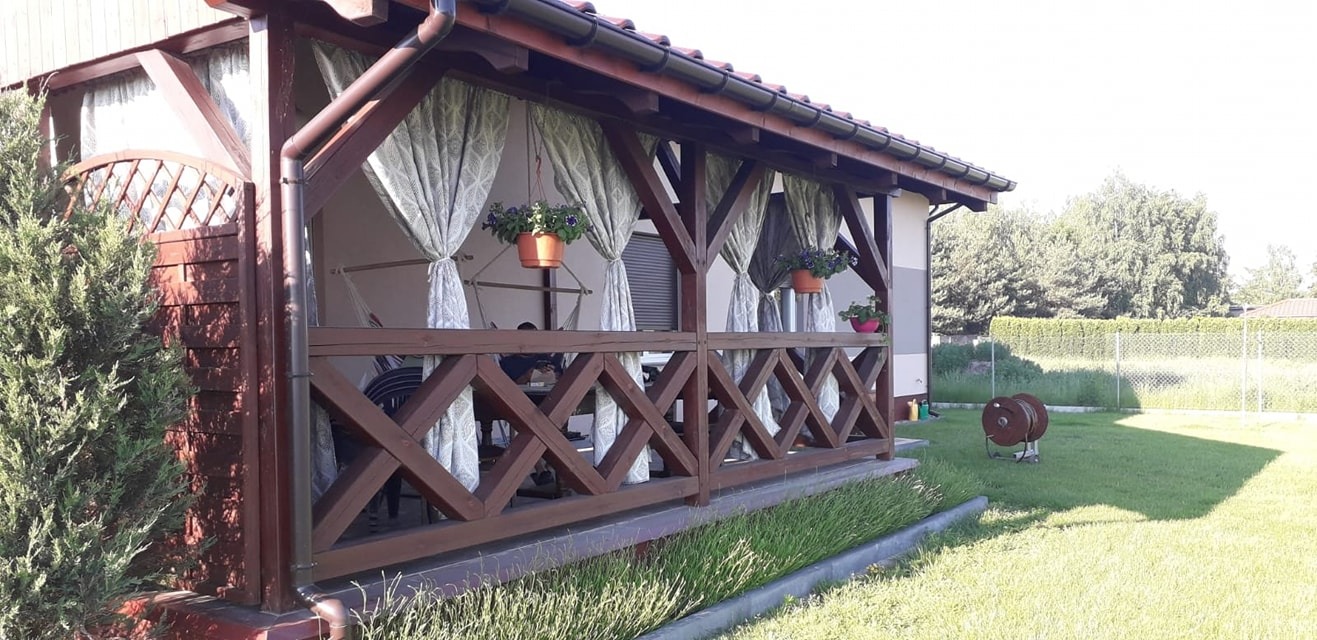 Drewniana altana ogrodowa z dekoracyjnymi zasłonami, wiszącymi doniczkami z kwiatami i kratownicą, otoczona zielenią.