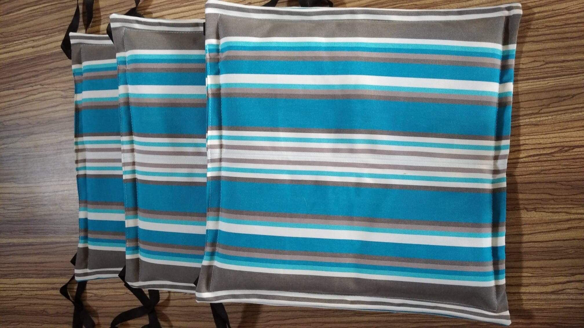 Trzy kwadratowe poduszki na krzesła z niebiesko-biało-szarymi paskami, z czarnymi troczkami do wiązania, ułożone na drewnianym tle.