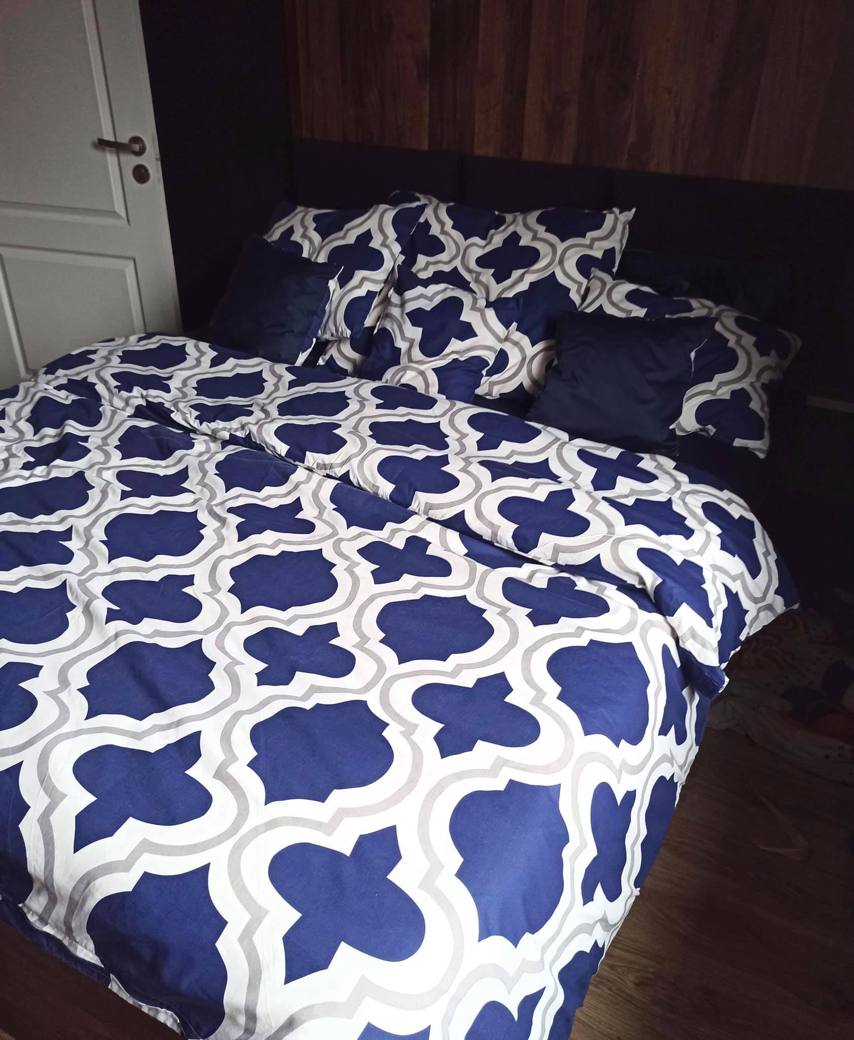 Gotowy komplet pościeli w odcieniach bieli, szarości i granatu, z geometrycznym wzorem marokańskim, ułożony na łóżku z ciemnym zagłówkiem i drewnianą podłogą, widoczna biała ściana z drzwiami.